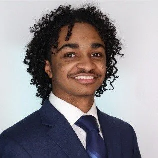 A young man with curly hair wearing a navy suit, white shirt, and navy tie, smiling and posing against a light background.