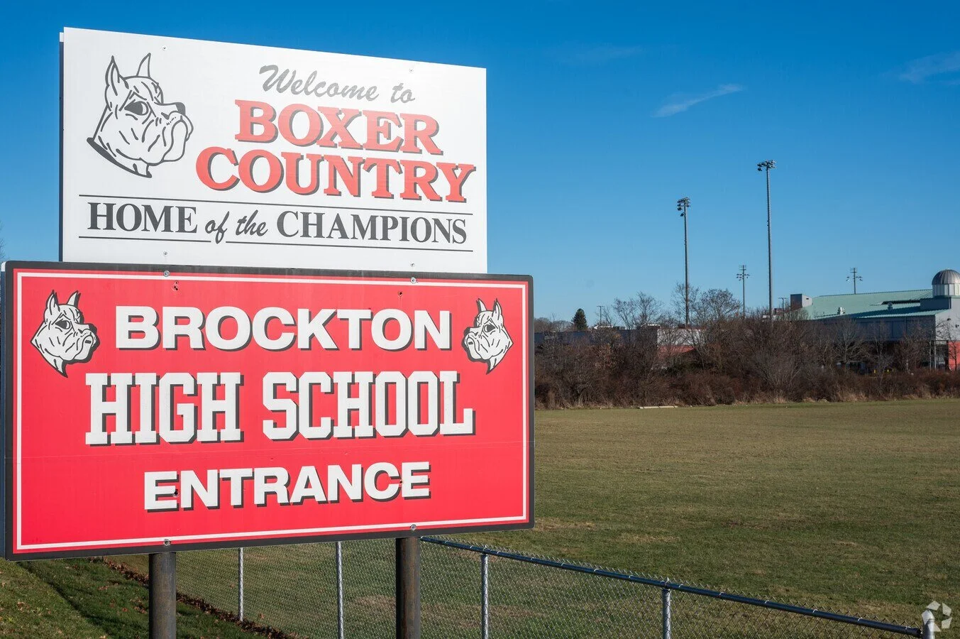 Signs indicating the entrance to Brockton High School and Boxer Country, with a field and school building in the background under a clear blue sky.