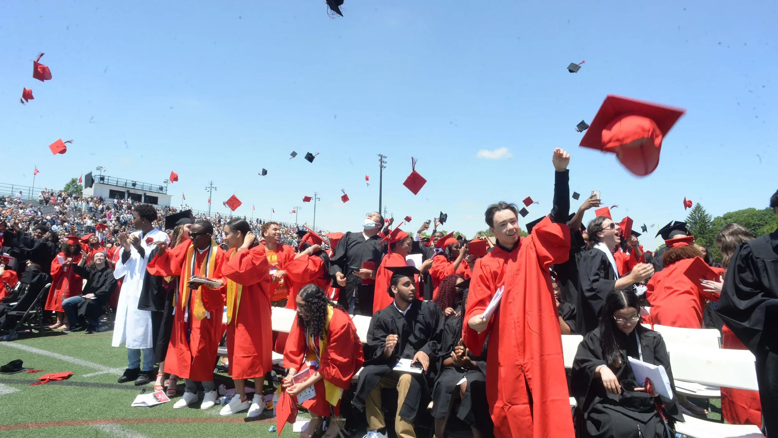 A large group of graduates in caps and gowns celebrating at a graduation ceremony outdoors, with many tossing their caps into the air.