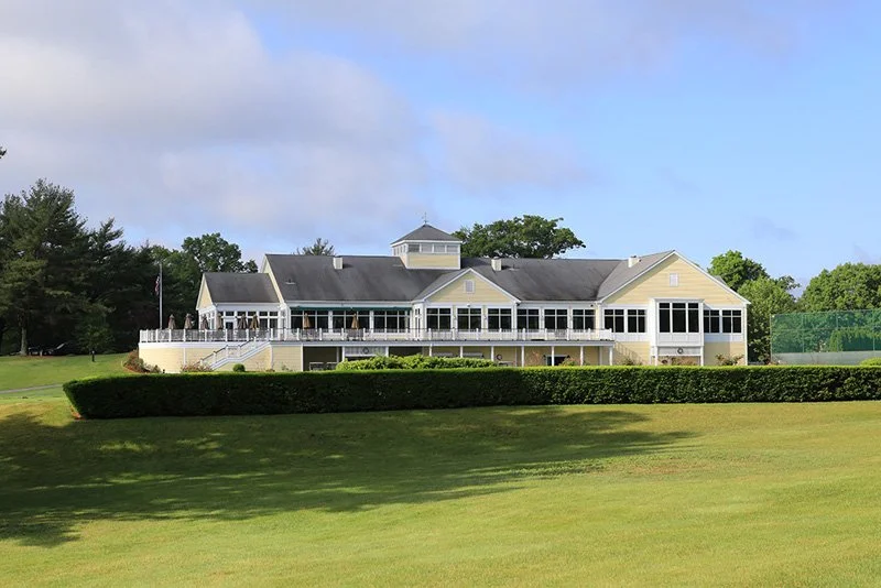 Large yellow clubhouse with windows and a wraparound porch, surrounded by a golf course with green grass and trees in the background.