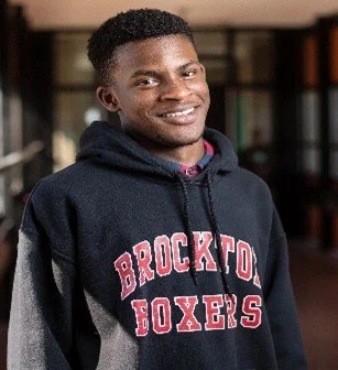 Young man smiling wearing a black hoodie with 'Brockton Boxers' in red letters, standing indoors.