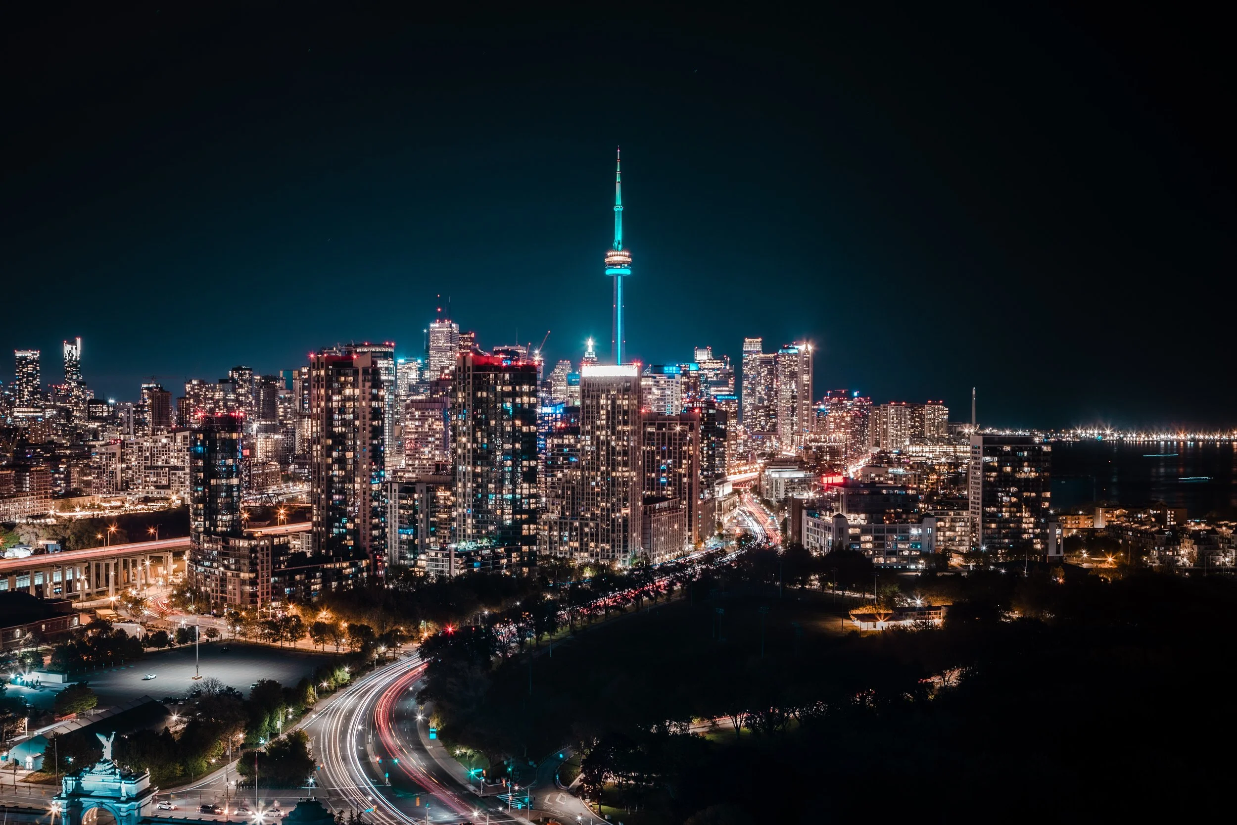 Nighttime cityscape of downtown Toronto, featuring the CN Tower illuminated in blue and surrounded by lit skyscrapers, with light trails of moving vehicles on the streets.