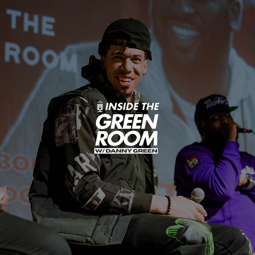 Young man with dark hair, wearing a black cap backwards and a camouflage jacket, smiling and holding a microphone, seated on stage. In the background, a woman in a purple hoodie holds a microphone, with a large projection of a man's face behind them.