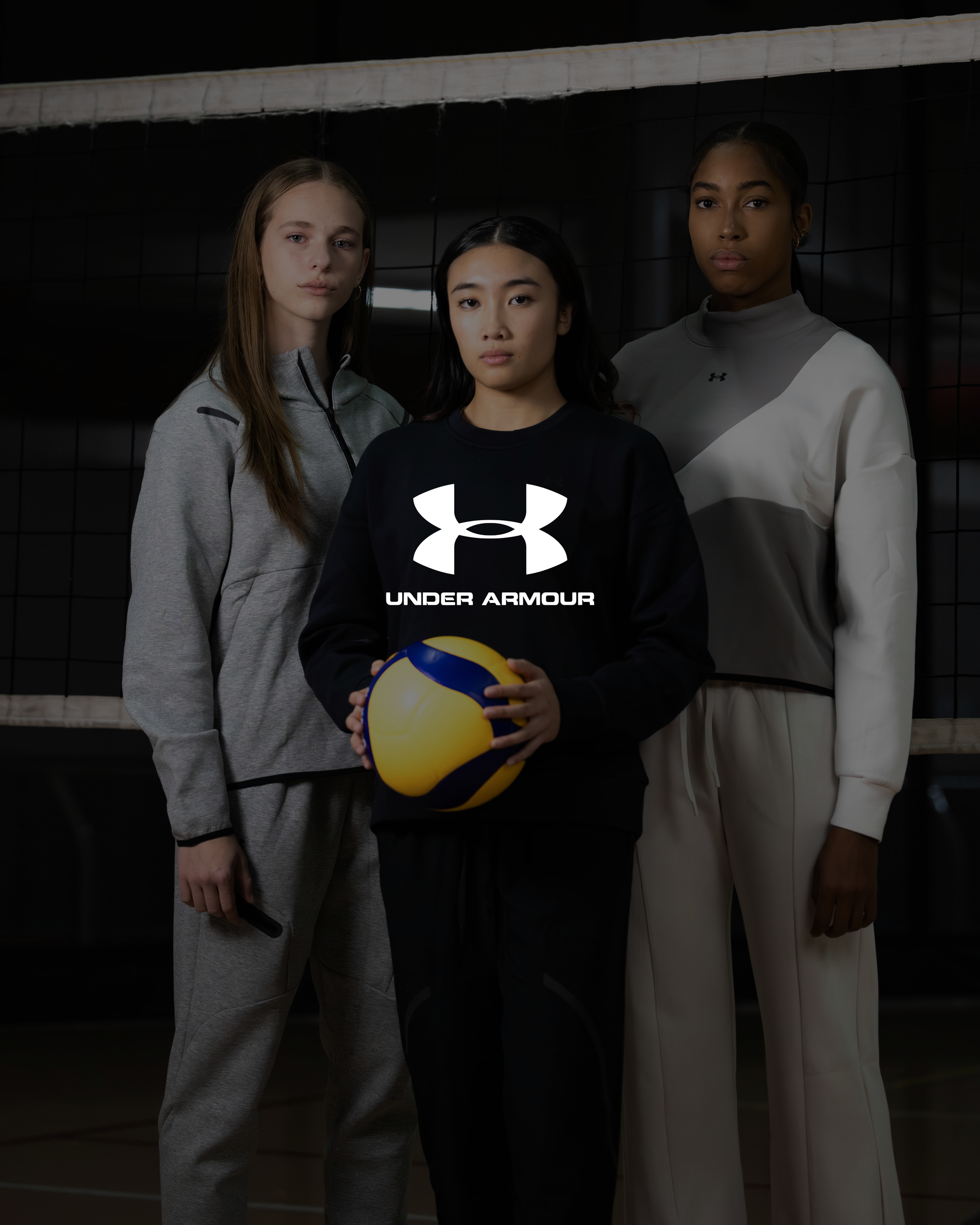 Three young women standing in front of a volleyball net in sportswear, with one holding a volleyball. The background is dark, and the women are looking directly at the camera.