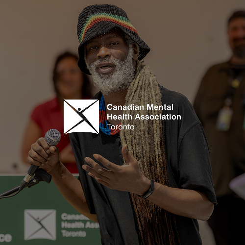 A man with gray hair and a beard, wearing a colorful hat and a scarf, speaking into a microphone at a Canadian Mental Health Association event in Toronto with two blurred people in the background.