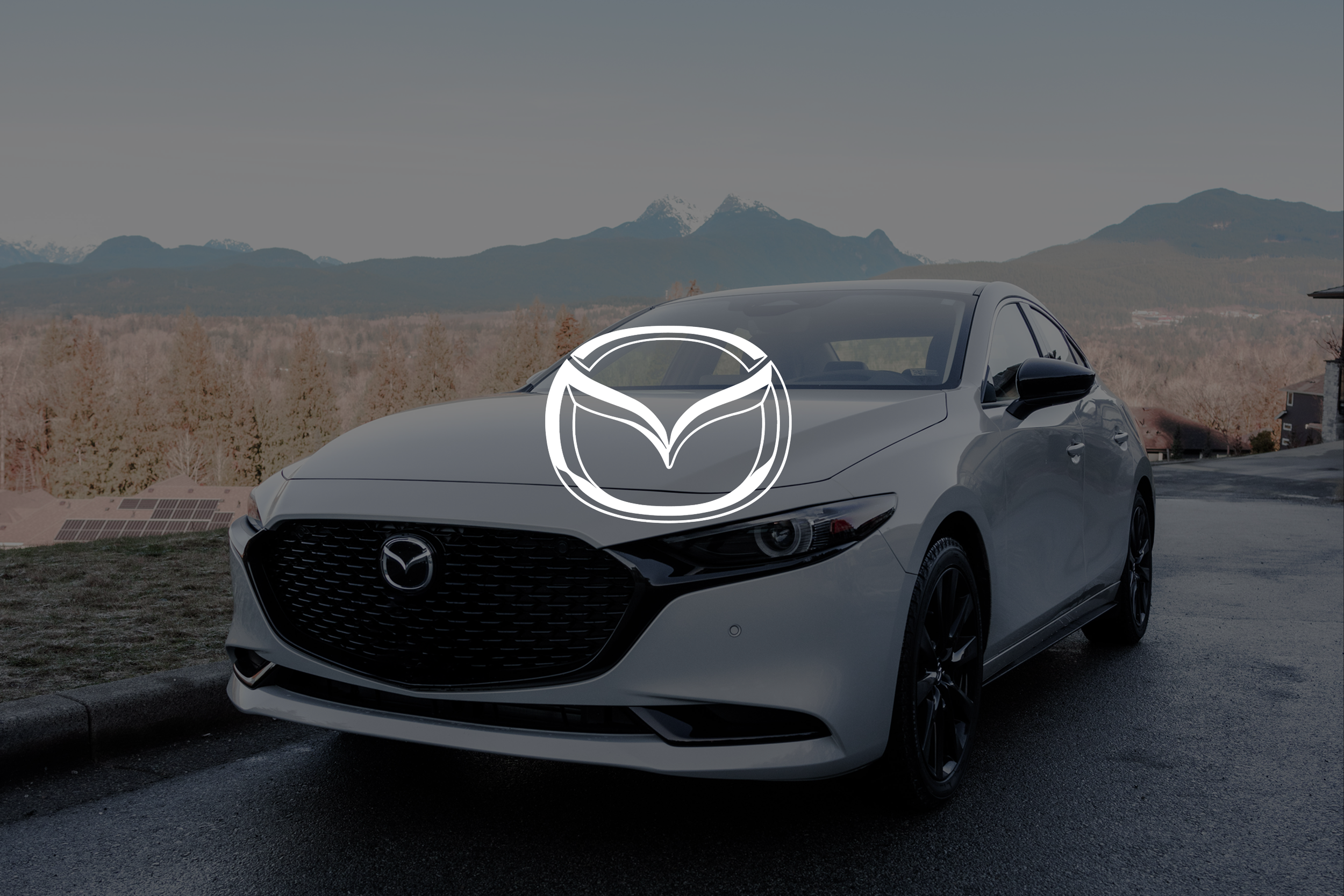 A silver Mazda sedan parked on a paved area with mountains in the background and a cloudy sky.