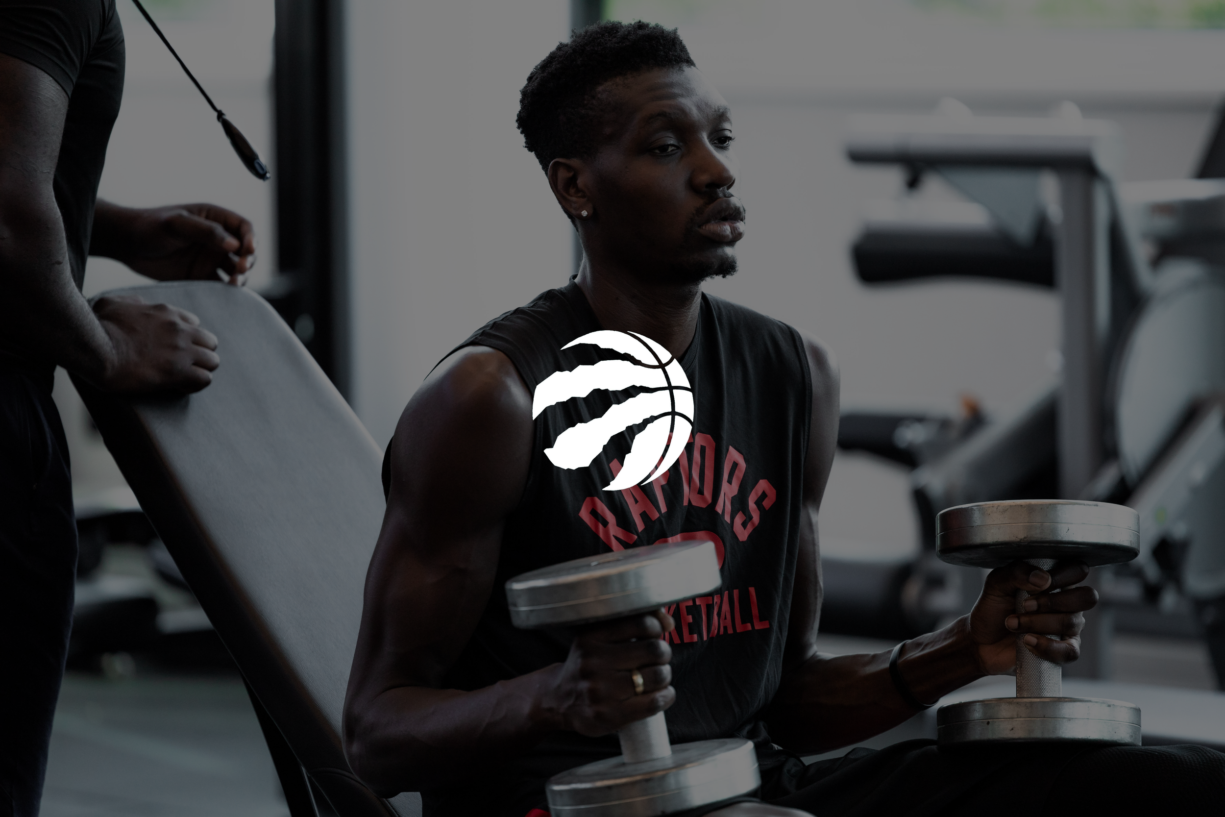 A man with dark skin and short hair working out with dumbbells in a gym, sitting on a weight bench next to a trainer.