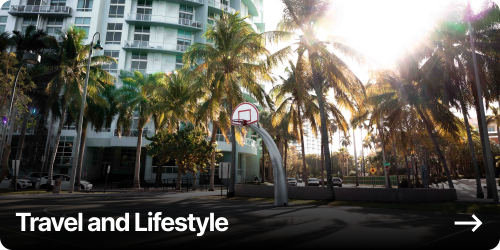 A city street with tall palm trees, a modern apartment building, and a basketball hoop in the foreground, illuminated by sunlight.