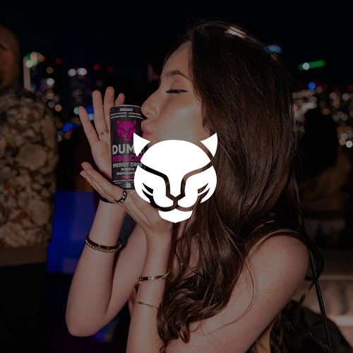 A woman with long dark hair closed eyes, holding a can of energy drink near her face at a nighttime event with city lights in the background.