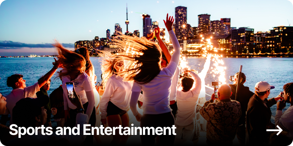 People dancing and celebrating near the water with fireworks and a city skyline in the background during evening.