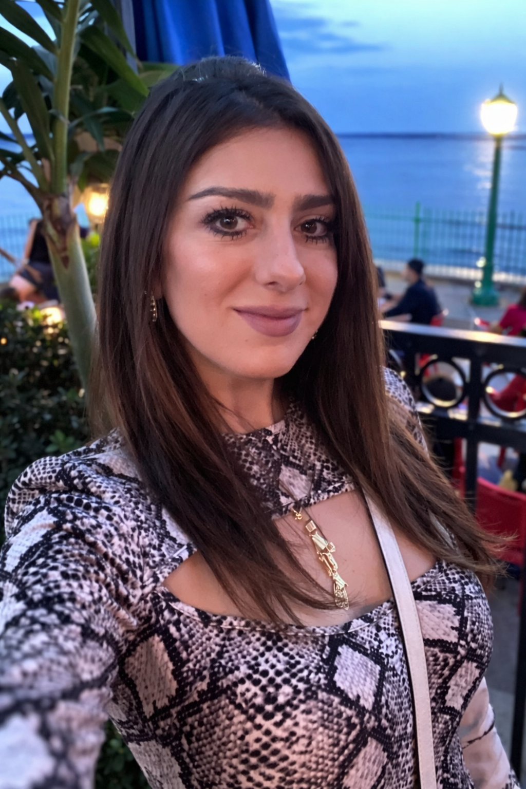 A young woman taking a selfie outdoors near a body of water during dusk. She has long dark hair, makeup, and is wearing a patterned top with cutouts and a gold necklace. There are people sitting at tables and a lamppost in the background.