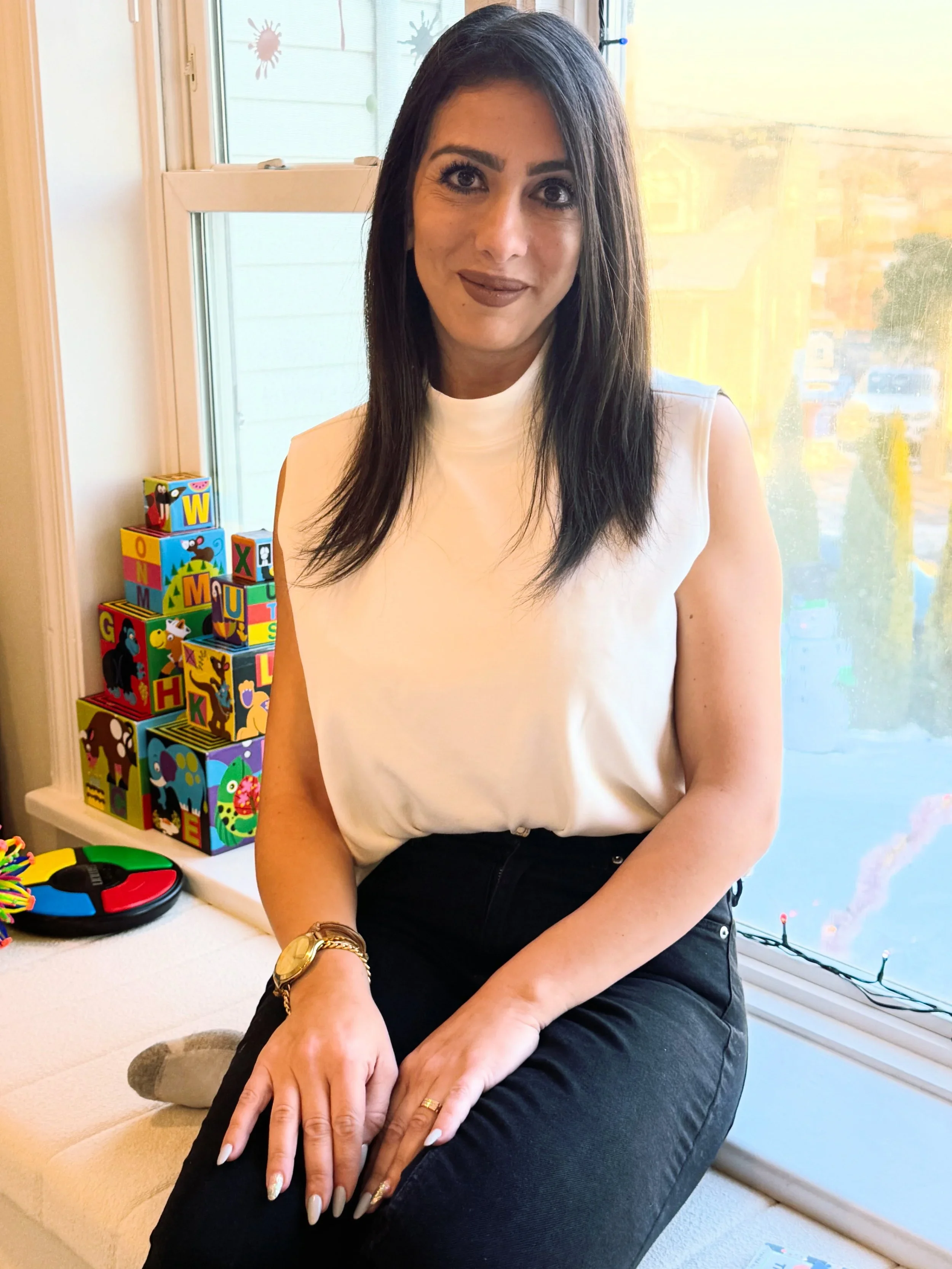 A woman with long dark hair, wearing a sleeveless white top and black pants, sitting on a beige surface near a window with colorful alpha blocks on a shelf nearby.