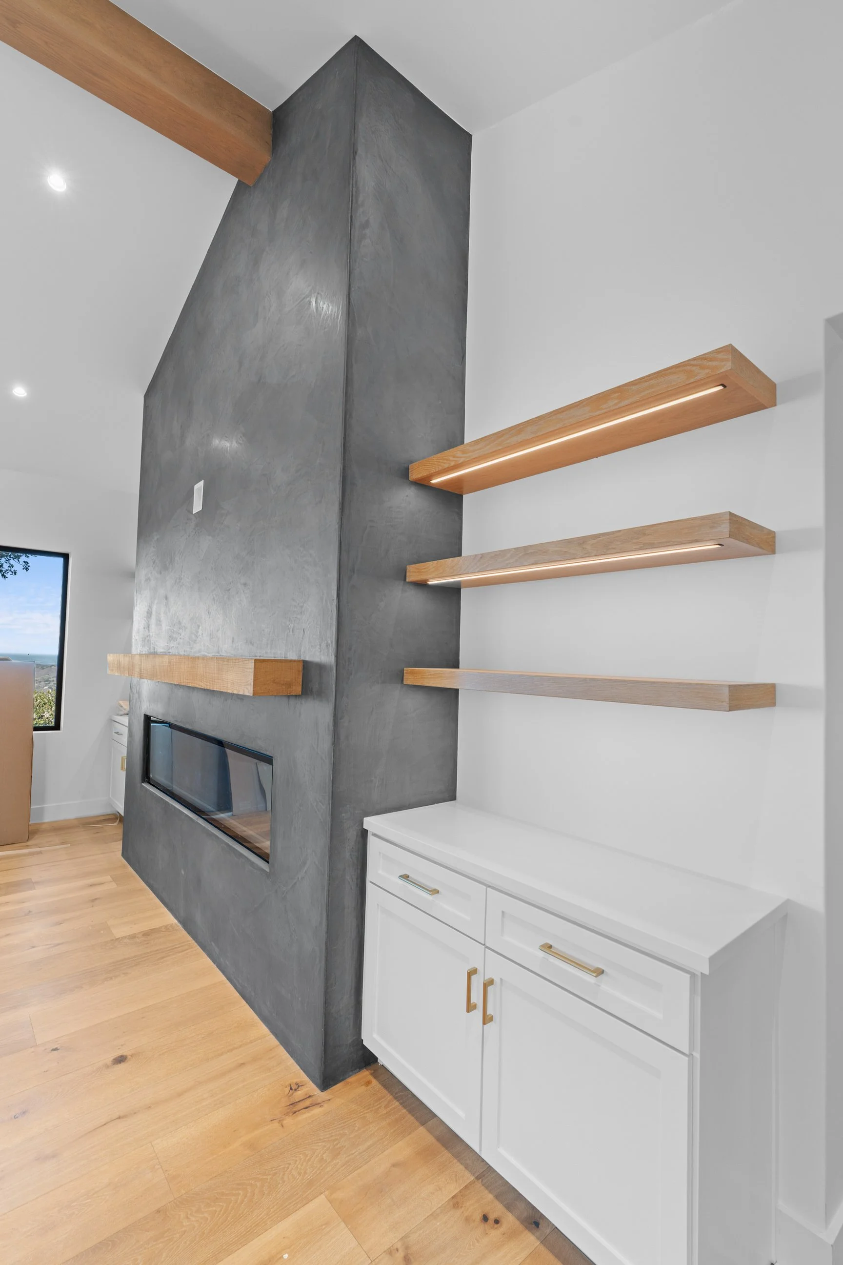 Interior view of a modern living room featuring a gray textured wall with a built-in fireplace, three floating wooden shelves with integrated lighting, white cabinetry with gold handles, hardwood flooring, and a window in the background showing a sunny outdoor scene.