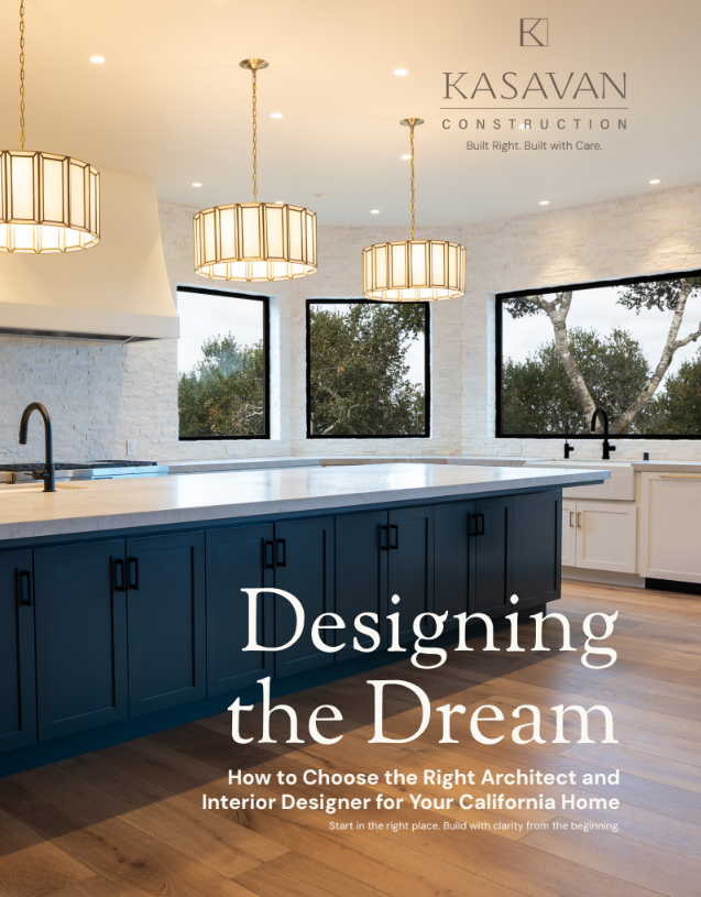Interior view of a modern kitchen with white brick walls, large black-framed windows showing trees outside, a navy blue kitchen island, and three pendant lights hanging from the ceiling. The cover features the title 'Designing the Dream' and the subtitle about choosing the right architect and interior designer for a California home, with the Kasavan Construction logo and slogan.