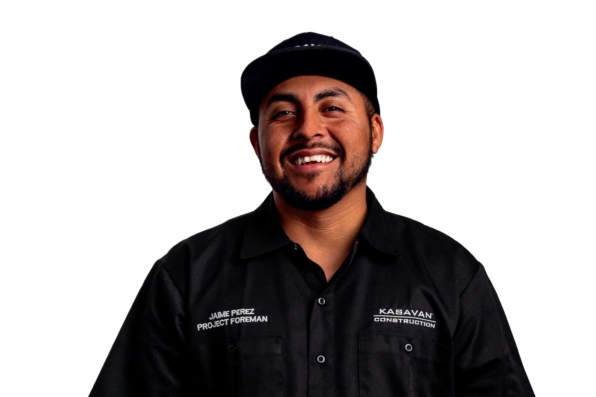 A man with a beard and mustache, smiling, wearing a black cap and a black shirt with white embroidered text.
