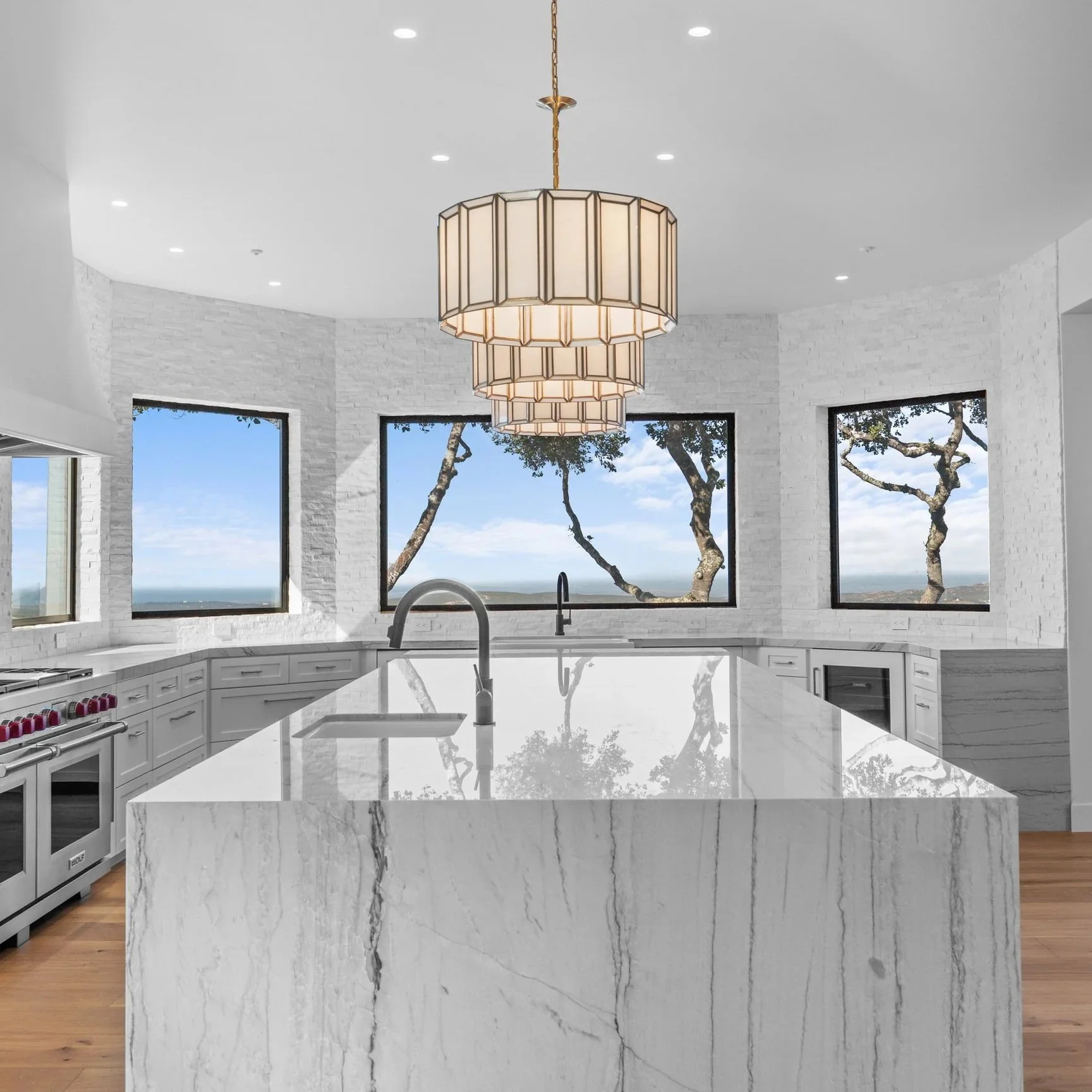 Modern kitchen with white marble island, white cabinets, large windows showing trees and blue sky, and pendant lights hanging from white ceiling.