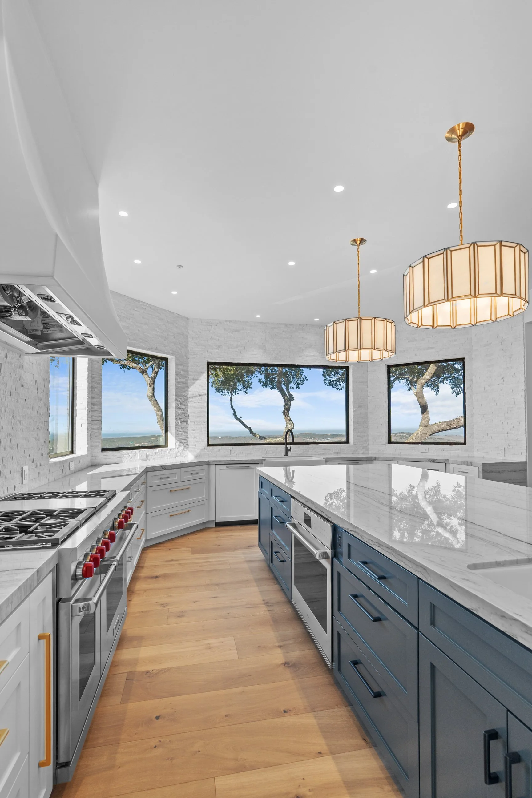 Modern kitchen with white walls and a marble island counter, blue lower cabinets, white upper cabinets, stainless steel appliances, wooden flooring, large windows with scenic views, and two pendant lights hanging from the ceiling.