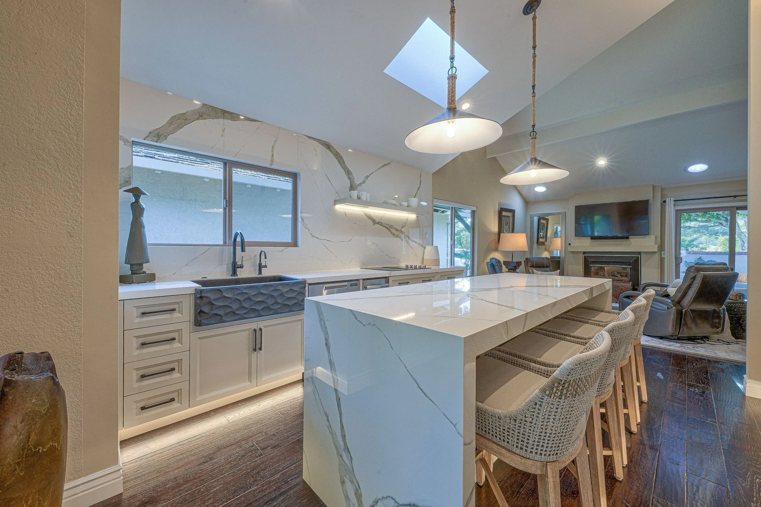 Modern kitchen and living room with marble island, bar stools, hanging pendant lights, fireplace, large flat-screen TV, and sliding glass doors with windows overlooking the outdoors.