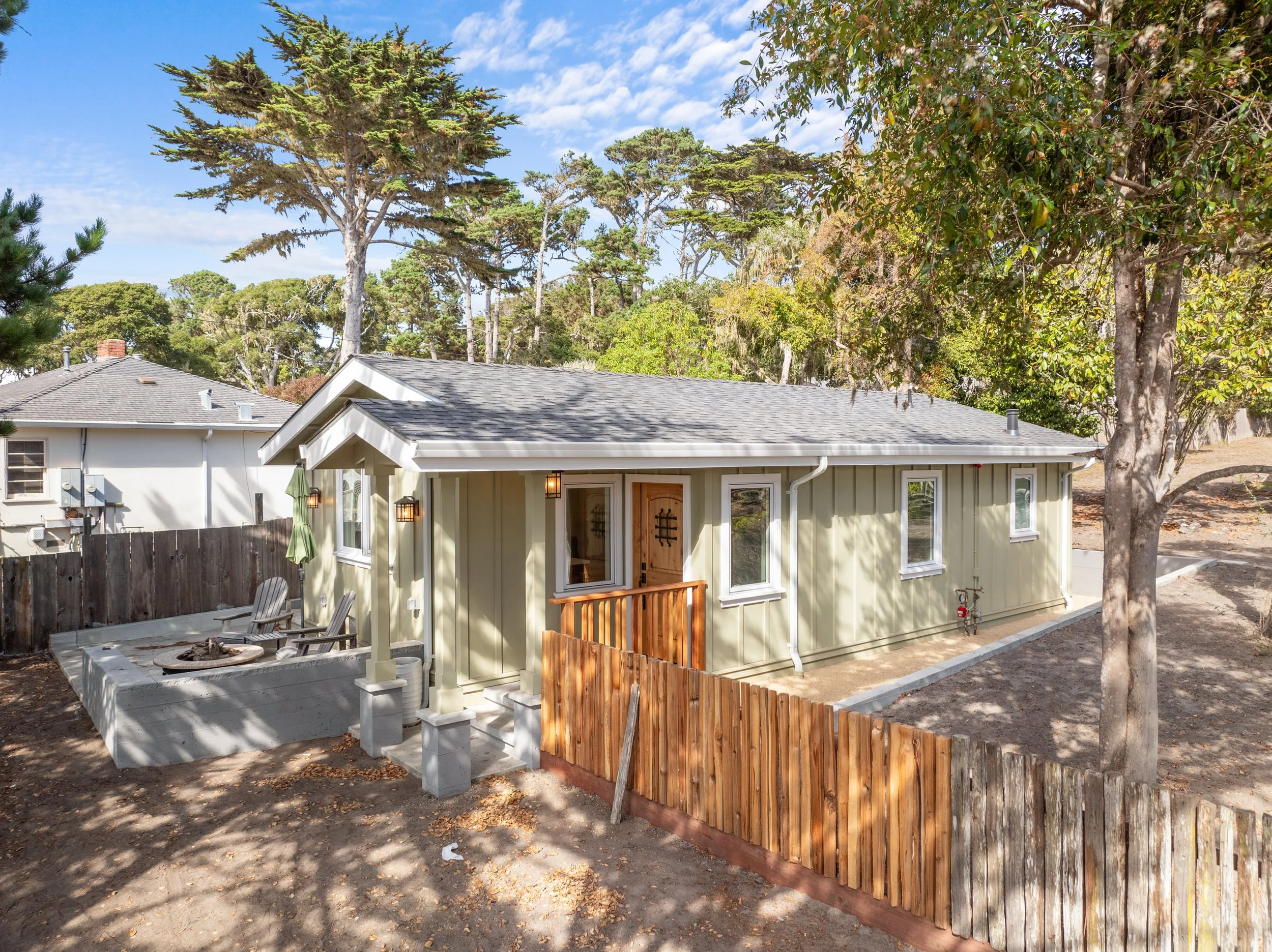 A backyard with a yellow house, large trees, a wooden fence, and outdoor seating with an umbrella.
