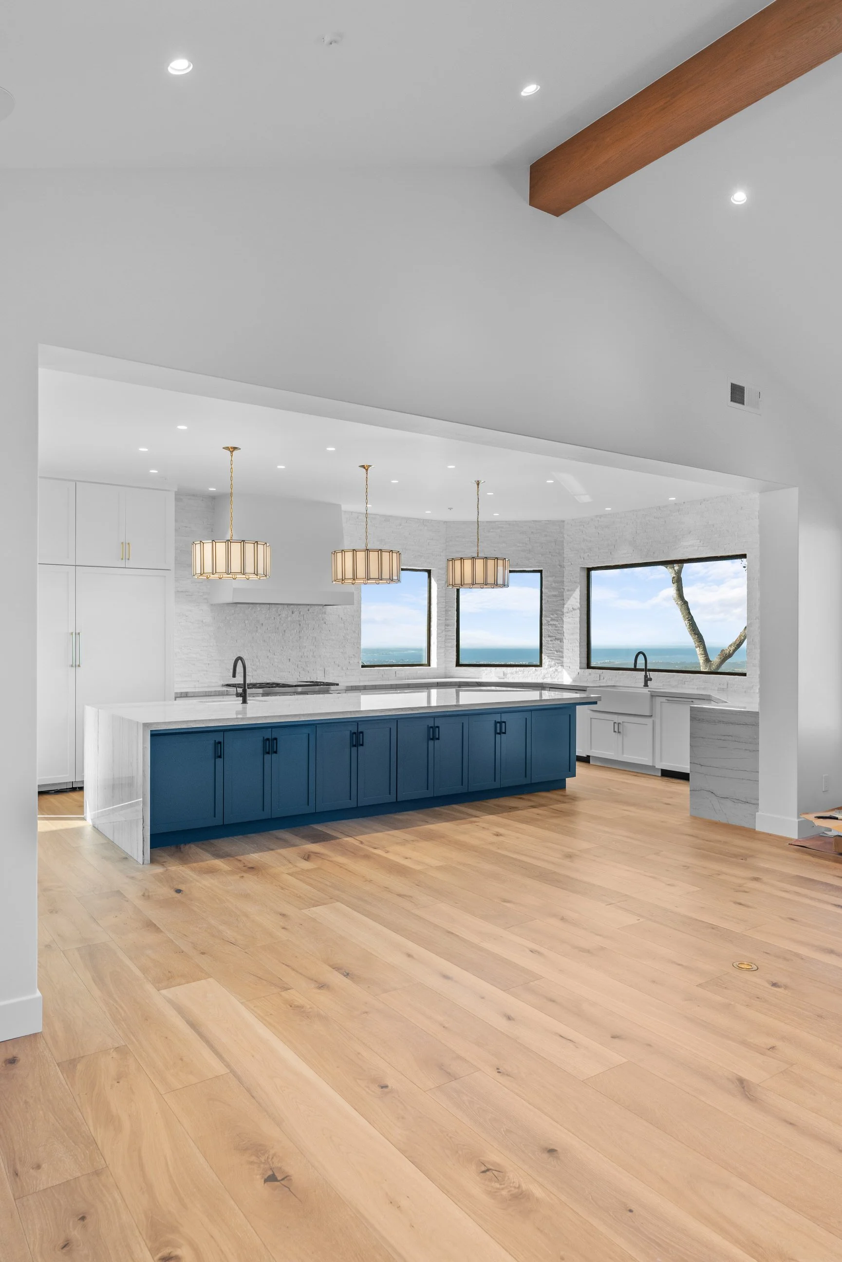 Modern kitchen with white cabinets, a large center island with a dark blue base, pendant lights, light wood flooring, large windows showing a tree and blue sky outside.