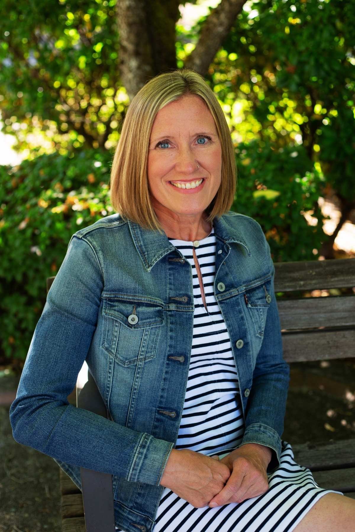 A woman with shoulder-length blonde hair, blue eyes, and a big smile sitting on a park bench. She is wearing a denim jacket over a navy and white striped dress, with a nose ring. The background shows green trees and sunlight filtering through the leaves.