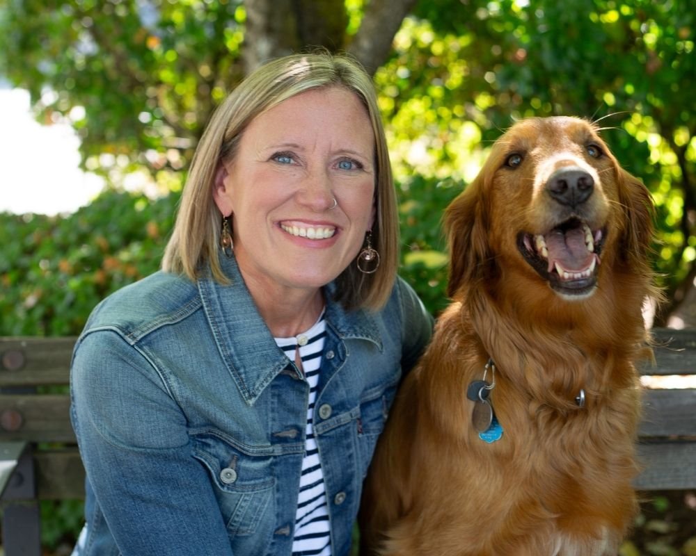 A smiling woman with blonde hair and blue eyes sitting outdoors next to a happy brown dog with a collar, in front of green trees.