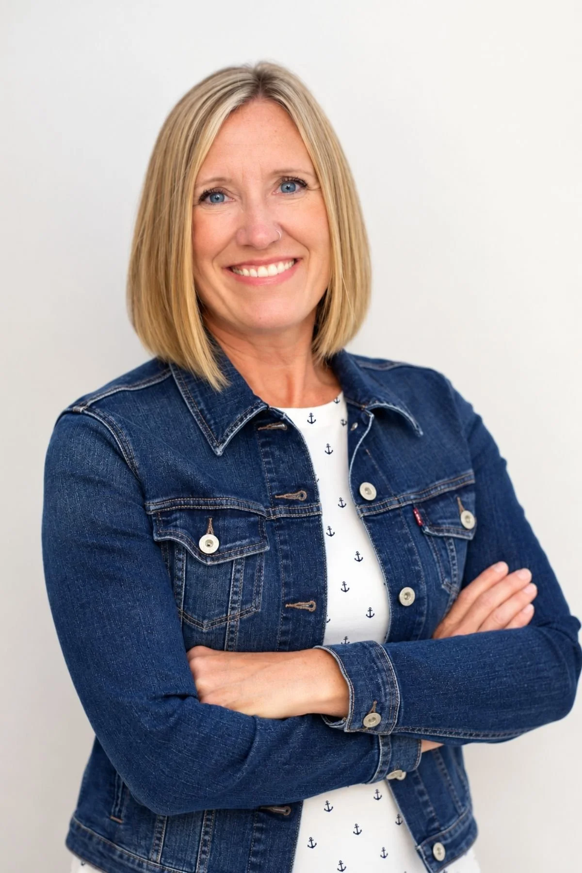 A woman with blonde hair, blue eyes, and a nose ring, smiling with crossed arms, wearing a denim jacket over a white dress with navy anchor prints. She is a licensed mental health counselor in Washington state.