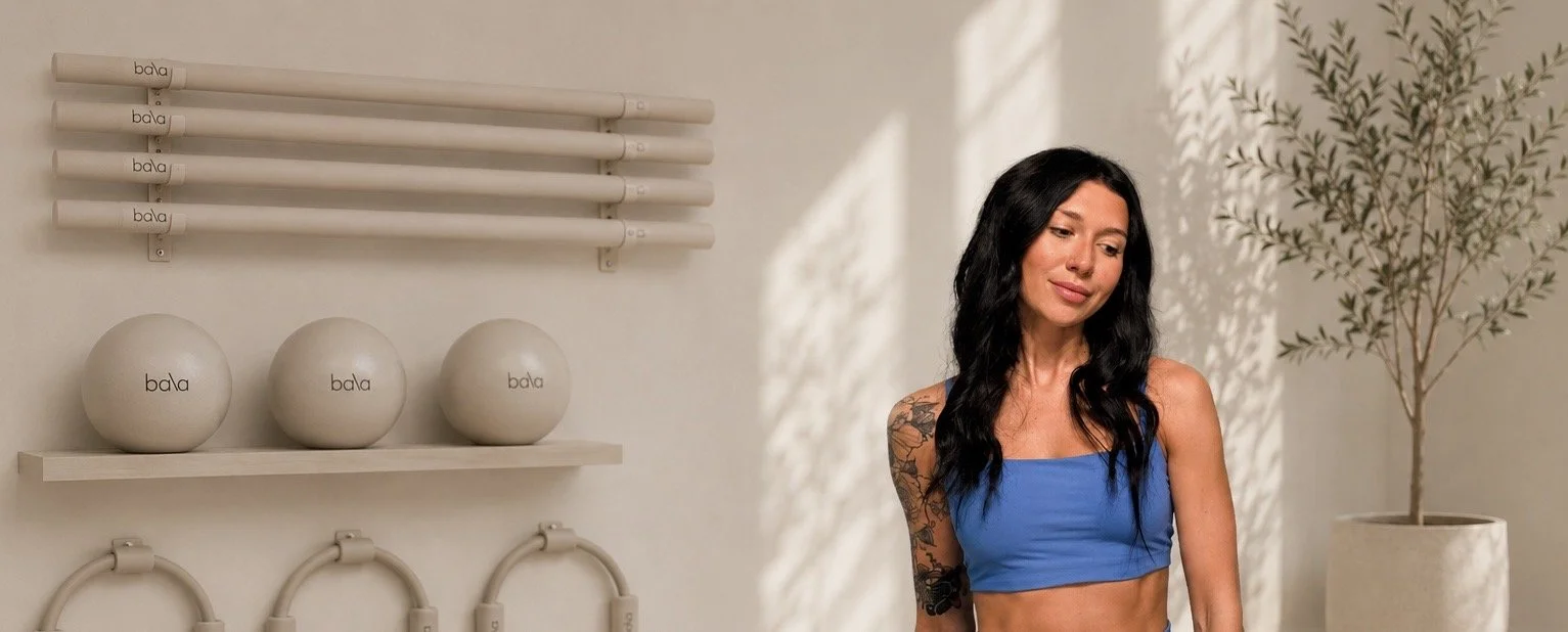 A woman with long black hair, wearing a blue sports bra, standing in a fitness studio with white walls, shelves holding exercise balls, and a potted plant.