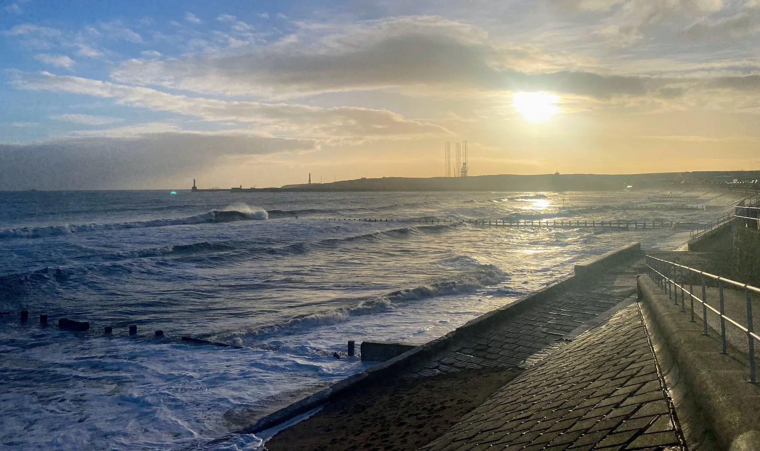 Sun setting over Aberdeen beach with waves crashing against the shore and an offshore drilling rig in the distance.