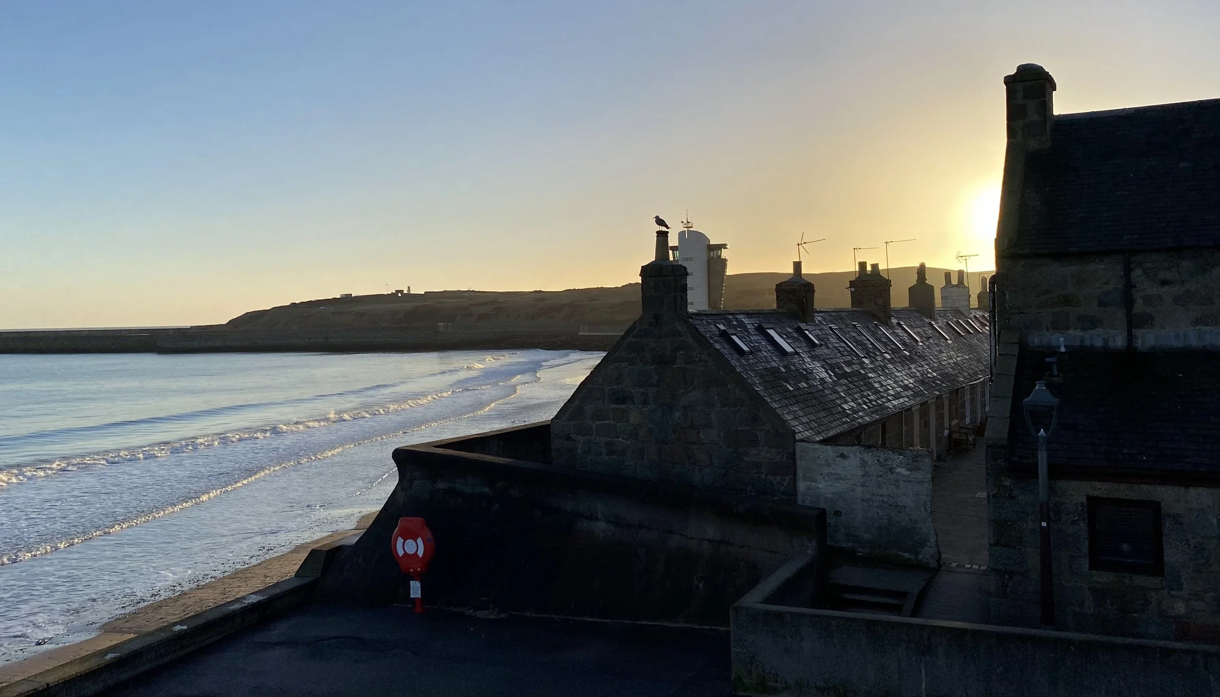 Sunset over a coastal village called Foot Dee ('Fittie') in Aberdeen with stone houses, chimneys, and rooftops, overlooking a beach with gentle waves. A seagull is perched on a chimney, and the sky is clear with the sun setting behind distant hills.