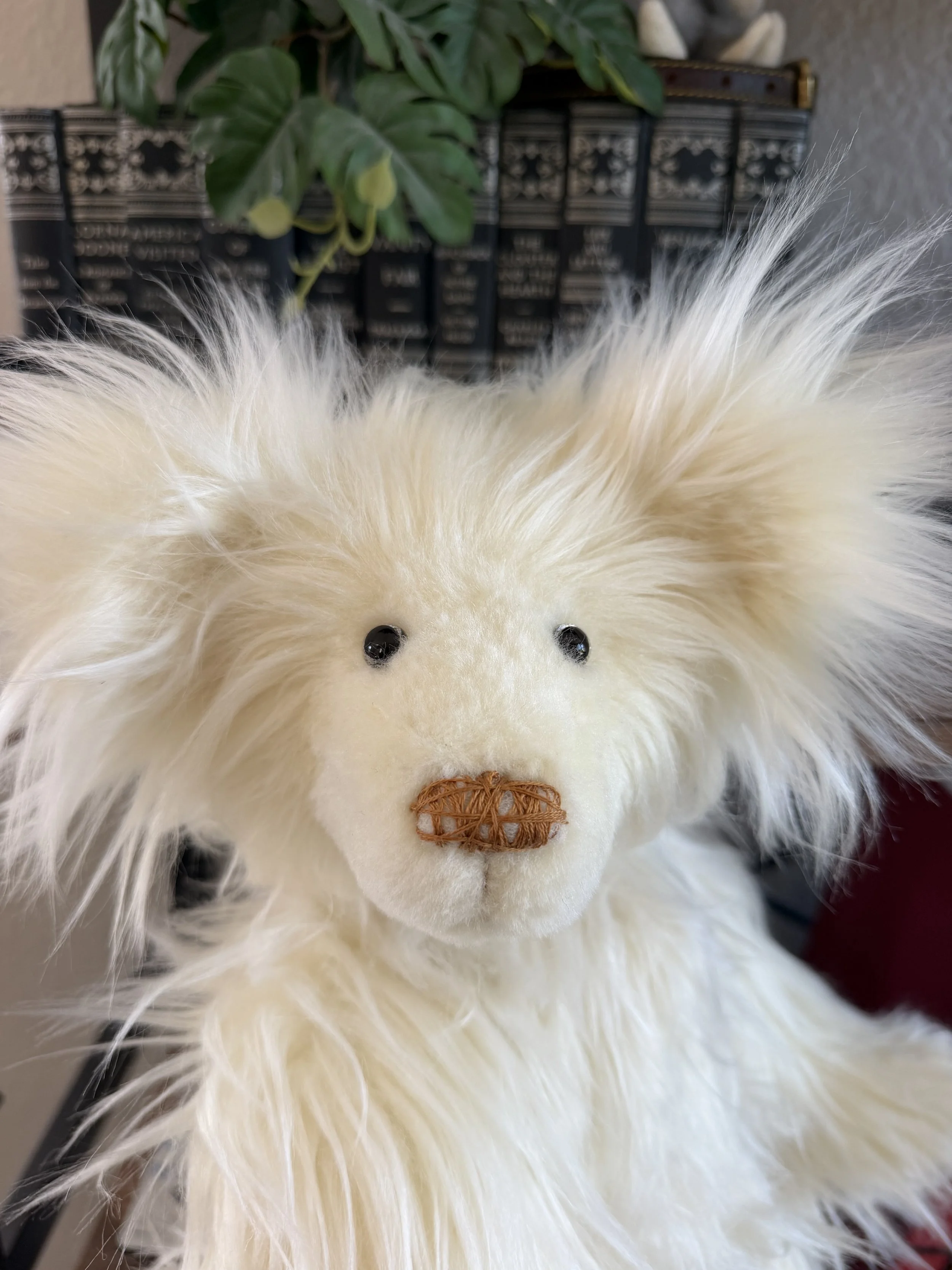 Close-up of a white plush hedgehog toy with black eyes and a stitched nose, positioned in front of a potted plant with large green leaves and a decorative black container with white patterns.