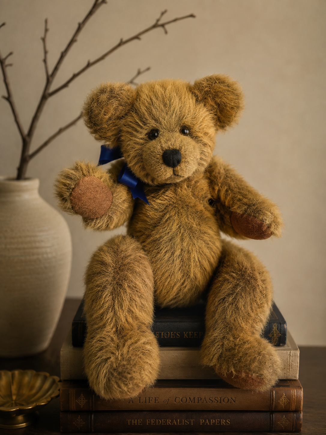 A teddy bear with a blue ribbon around its neck sitting on a stack of old books, with a vase and bare branches in the background.