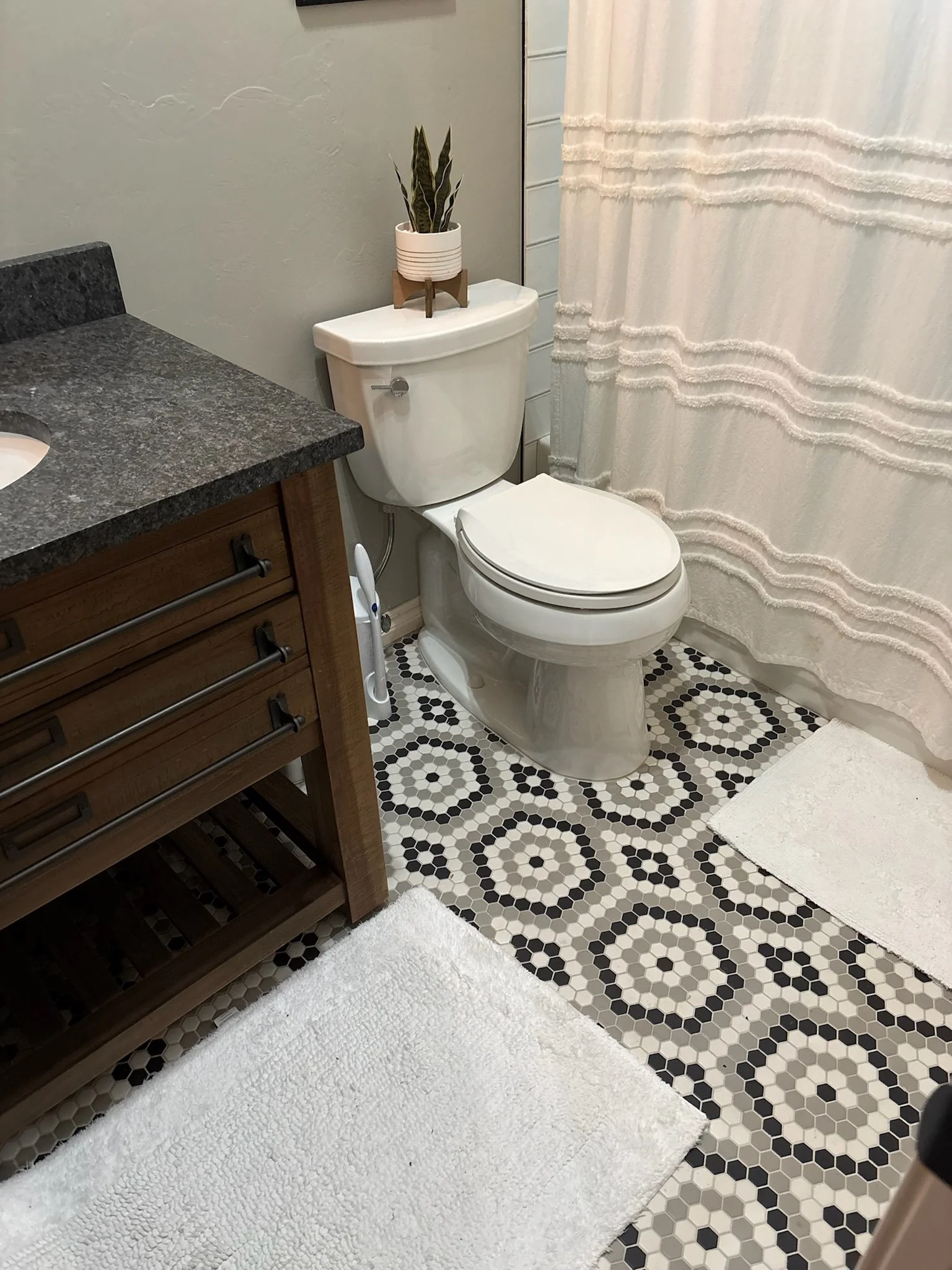A bathroom with a white toilet, a dark wooden vanity with a granite countertop, a plant in a white pot on top of the toilet tank, white fluffy rugs on a black and white patterned tile floor, and a white shower curtain.