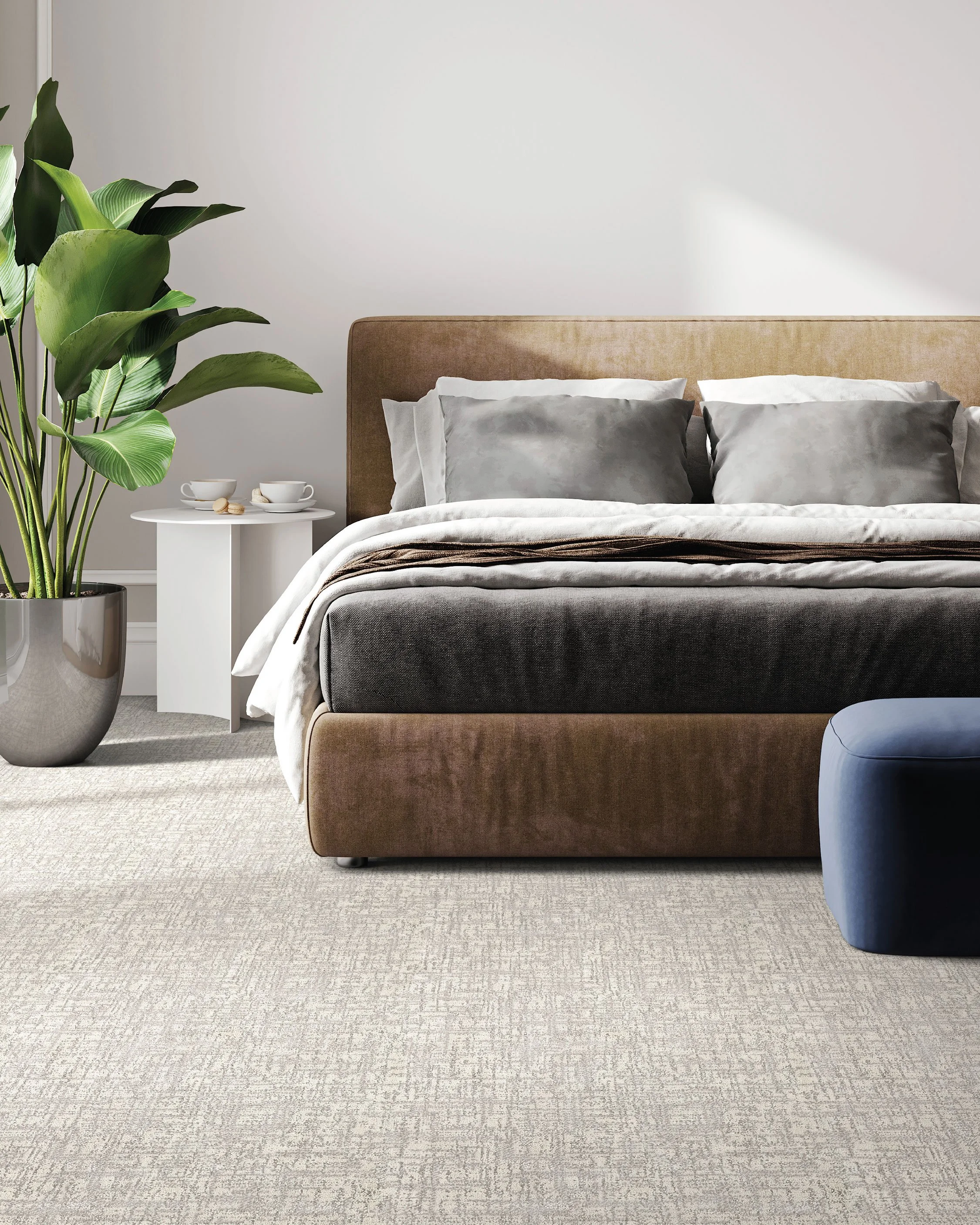 A neatly made bed with a beige headboard, gray and white pillows, and a white blanket, next to a white side table with two teacups and macarons. A large potted green plant is on the left, and a round blue stool is partially visible on the right, with a plain white wall in the background.