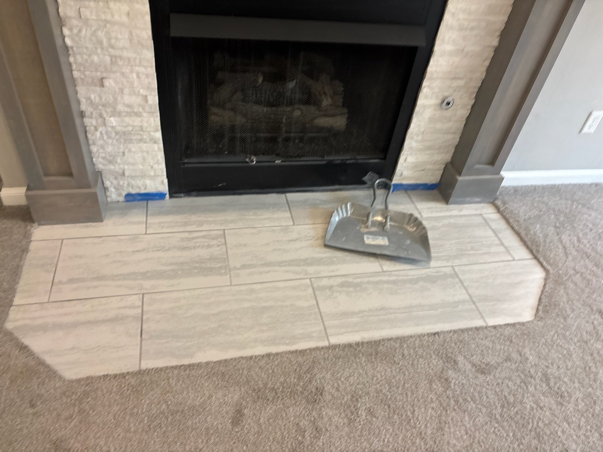 Fireplace with white brick surround, black metal frame, and logs inside, with a dustpan and broom in front and gray trim around the hearth.