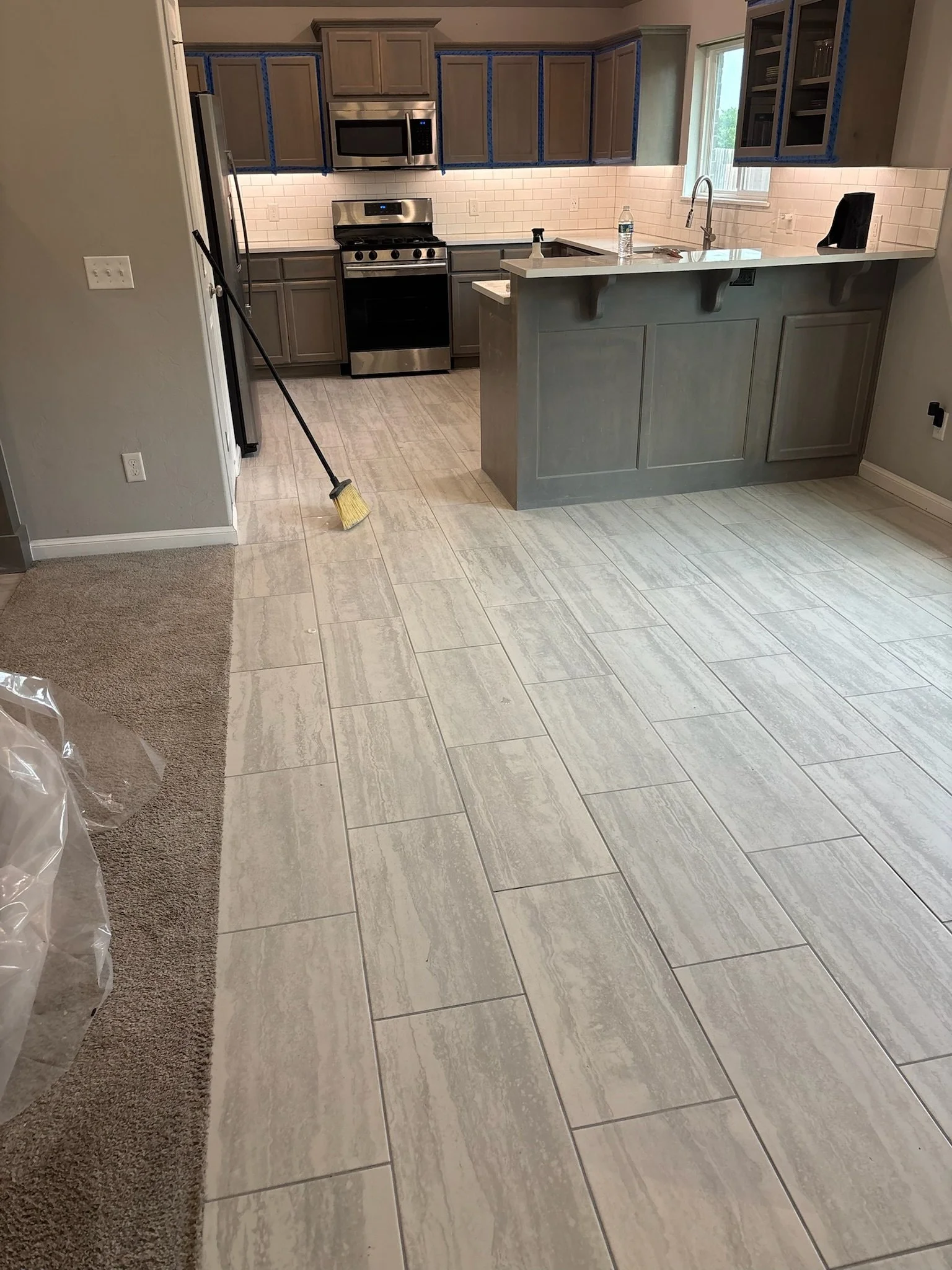 A modern kitchen with light-colored wood cabinets, stainless steel appliances, a tiled backsplash, a large window over the sink, and a countertop with a water bottle, a spray bottle, and a black speaker. The floor is tiled with large, beige tiles, an