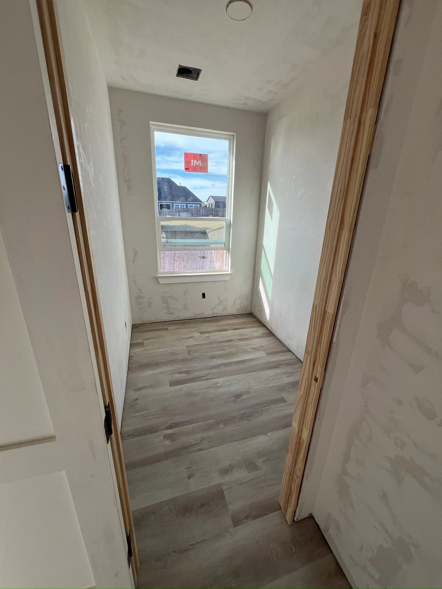 A small, unfinished room with a window, light-colored wood flooring, and walls painted white with some drywall patches. There is a ceiling light fixture and an air vent hole in the ceiling. The door frame is made of wood, and part of the door is visi