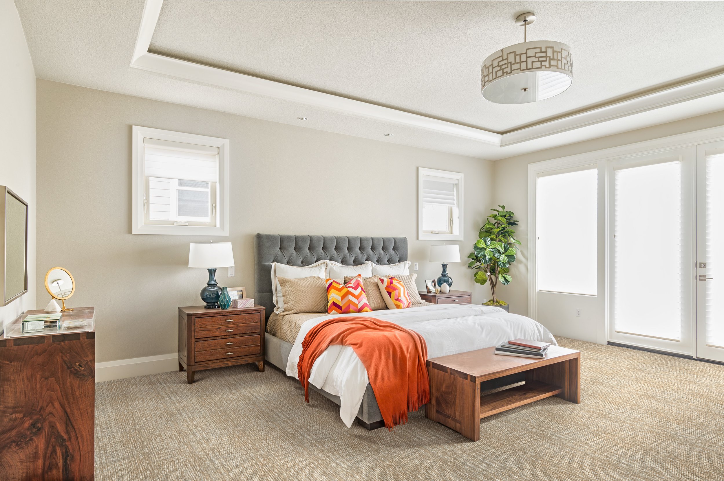 A bedroom with a large bed, a gray upholstered headboard, colorful pillows, an orange throw blanket, and two wooden nightstands with lamps. There are windows with white blinds, a potted plant, and a ceiling light fixture.