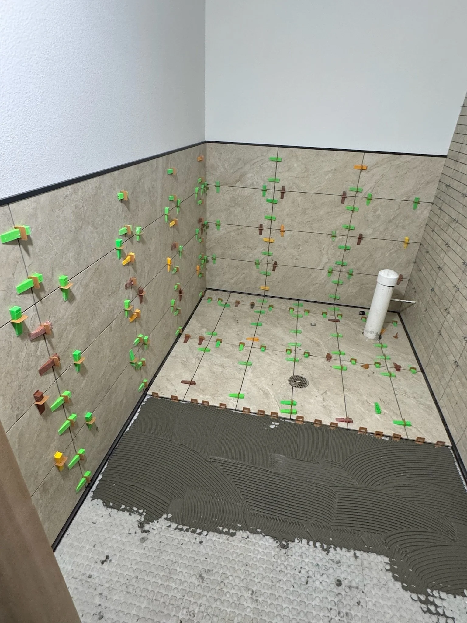 Bathroom wall tiling in progress with tiles held in place by colorful spacers, a drain, and a curved shower floor area with textured non-slip surface.
