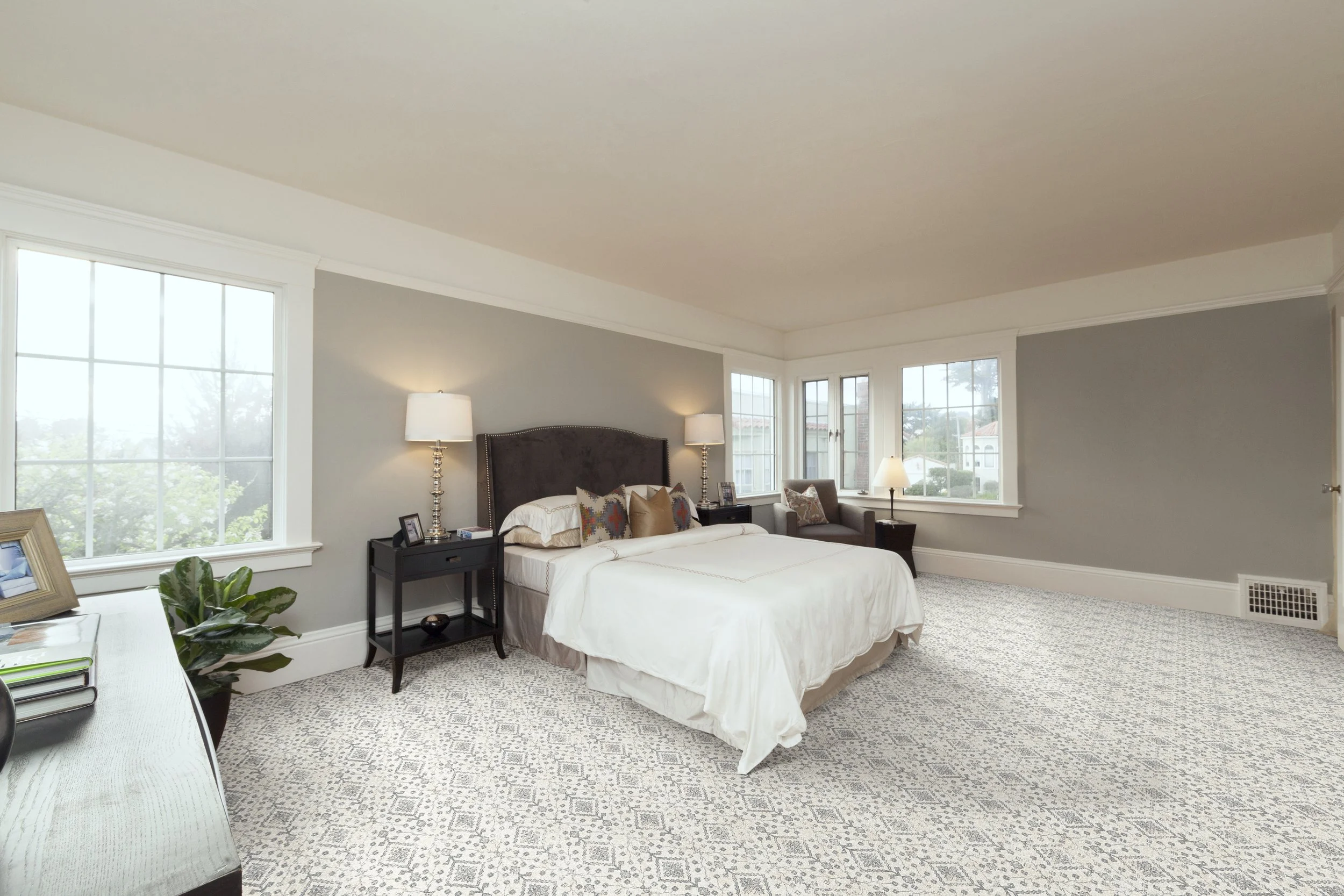 A spacious bedroom with large windows allowing natural light, a bed with decorative pillows and a dark headboard, two bedside lamps, a side table, a chair with pillows, and a patterned carpet.