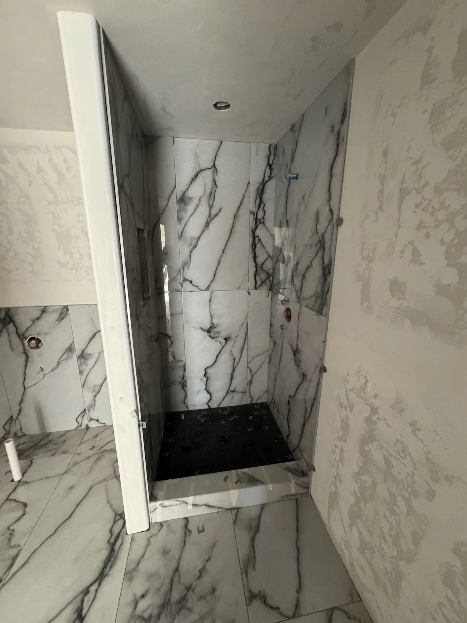 An unfinished bathroom with marble tiles on the walls and floor. There is a constructed shower with a black base. The ceiling shows an opening for a light fixture, and a strip of white material runs along the top edge of the marble walls. Constructio