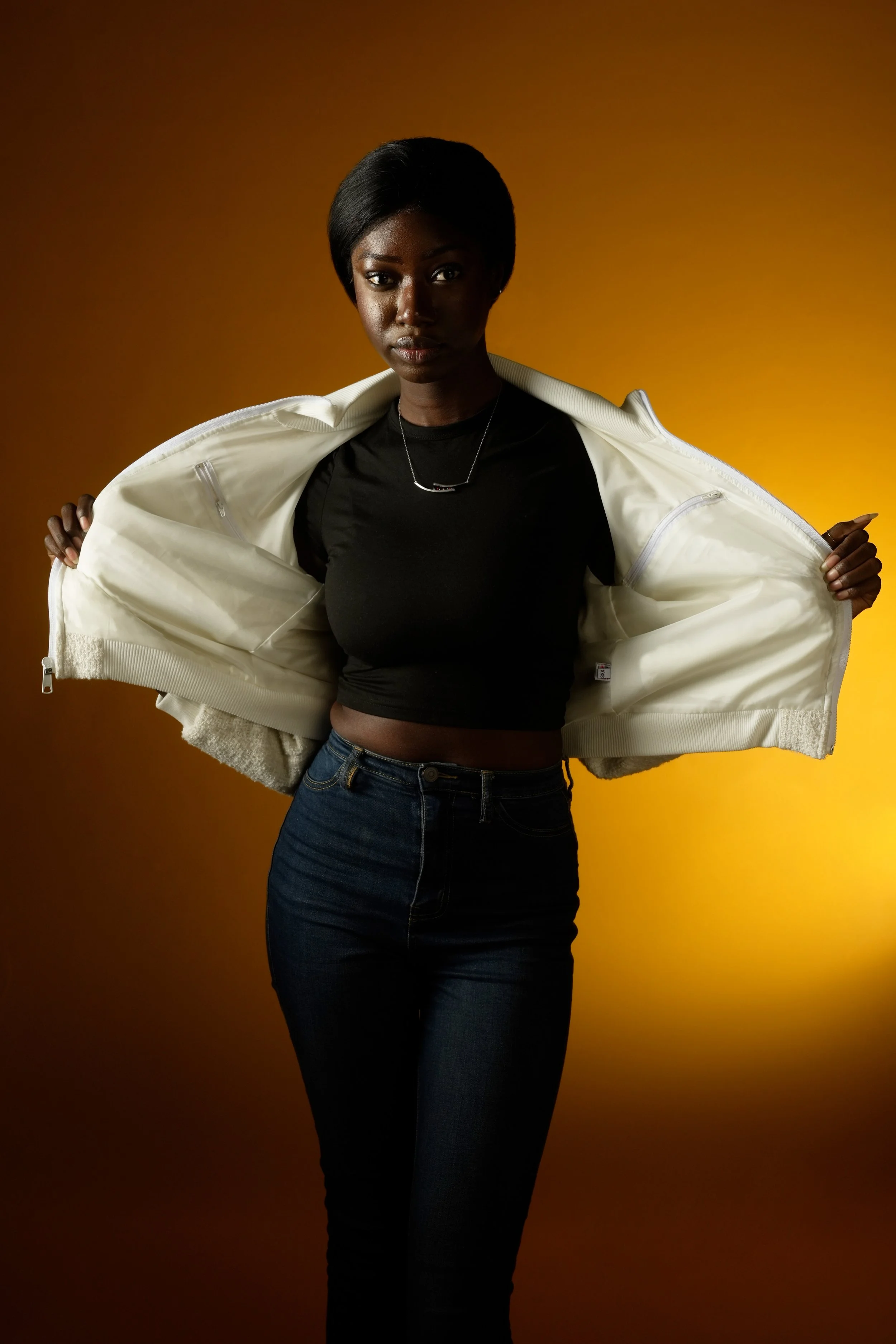 A woman with dark hair wearing a black top and dark jeans, holding open a white jacket against a warm background.