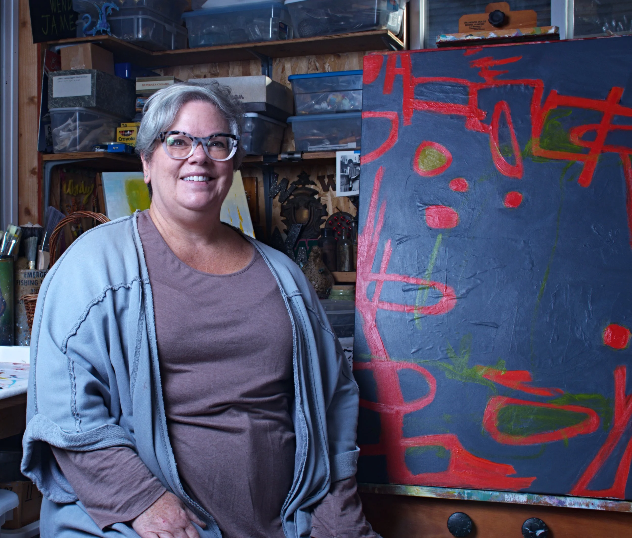 Smiling woman with short gray hair and glasses standing in front of a colorful abstract painting in a cluttered art studio.