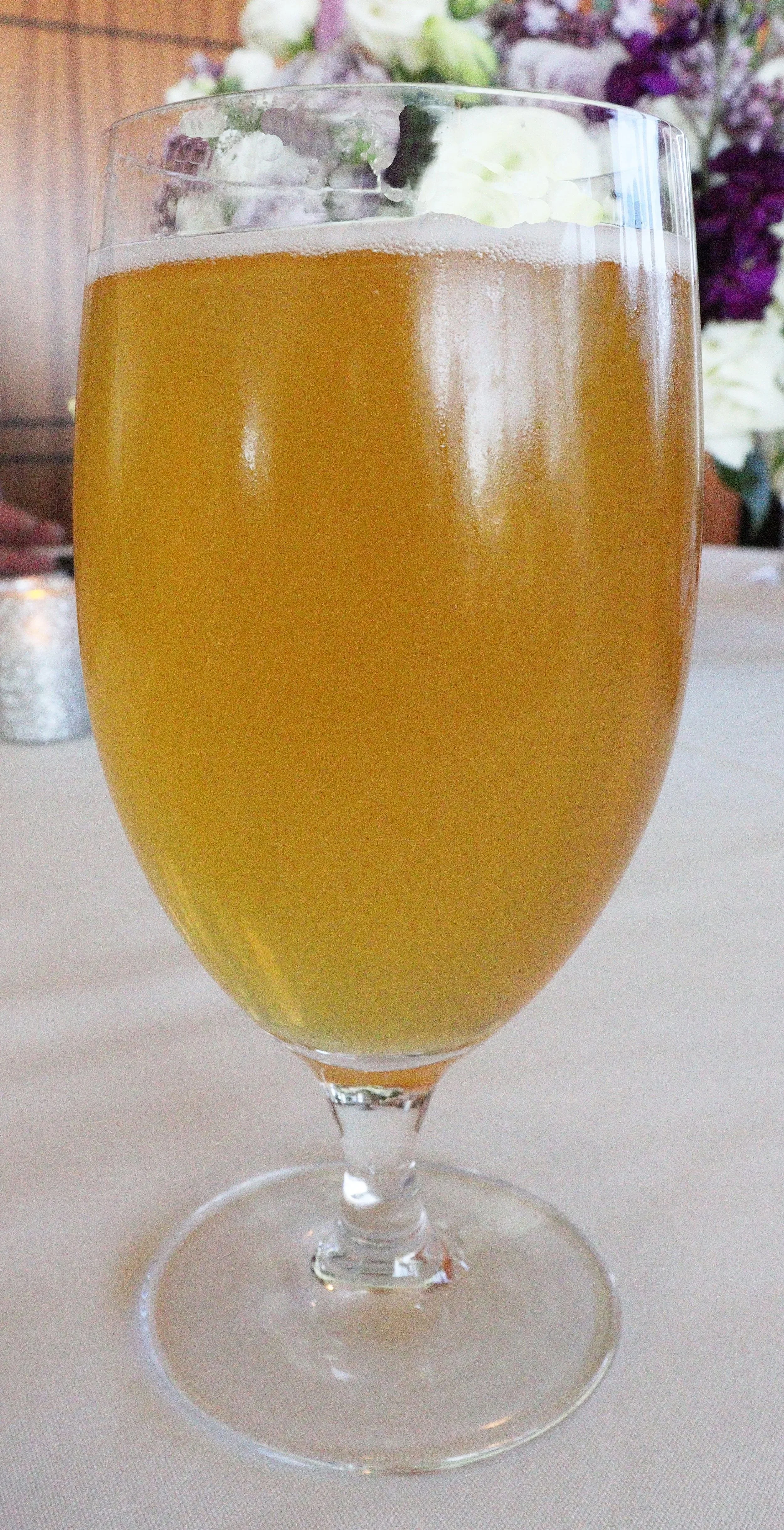 A tall glass of pale yellow, effervescent beverage, topped with a dollop of whipped cream, on a white table with flowers in the background.