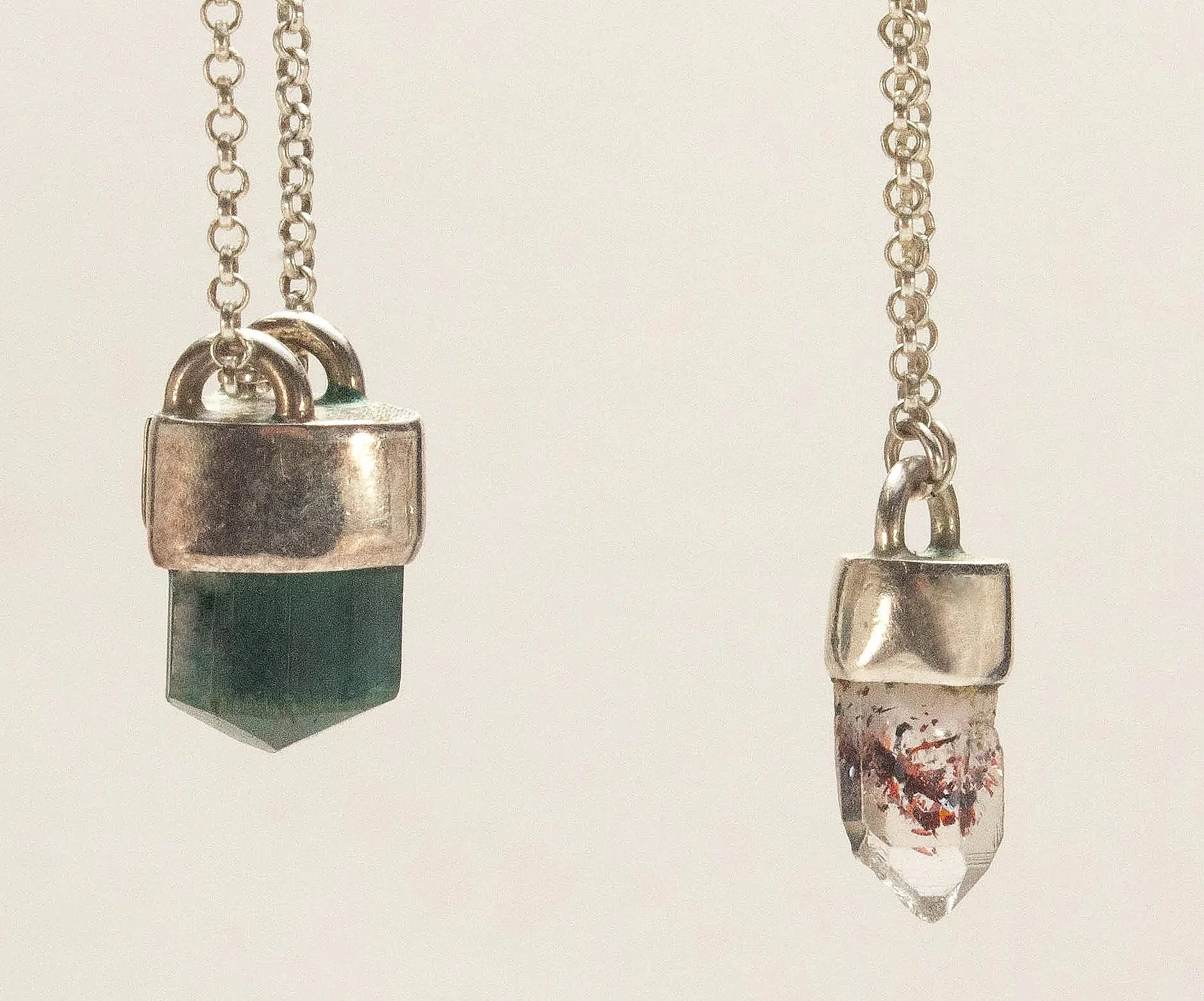 Close-up of a silver necklace with a cube-shaped pendant and a silver chain, alongside a painted glass stand with a small pointy pendant, both hanging against a plain white background.