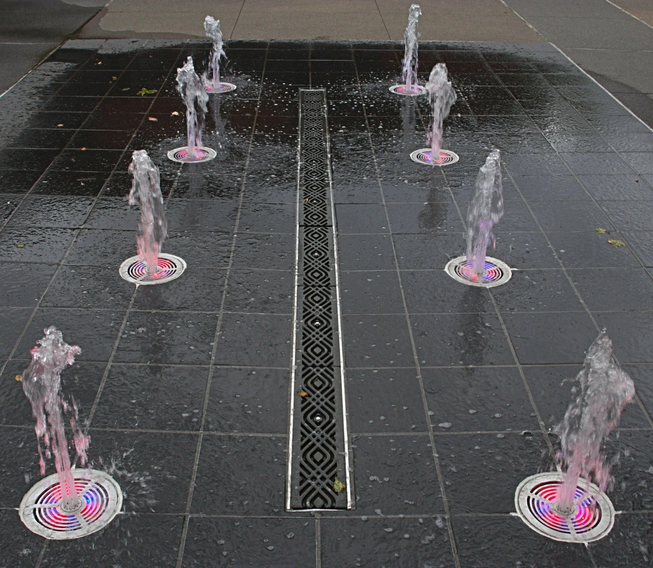 A small outdoor water fountain with multiple streams of water shooting upward through circular drainage holes in a wet black tiled surface.