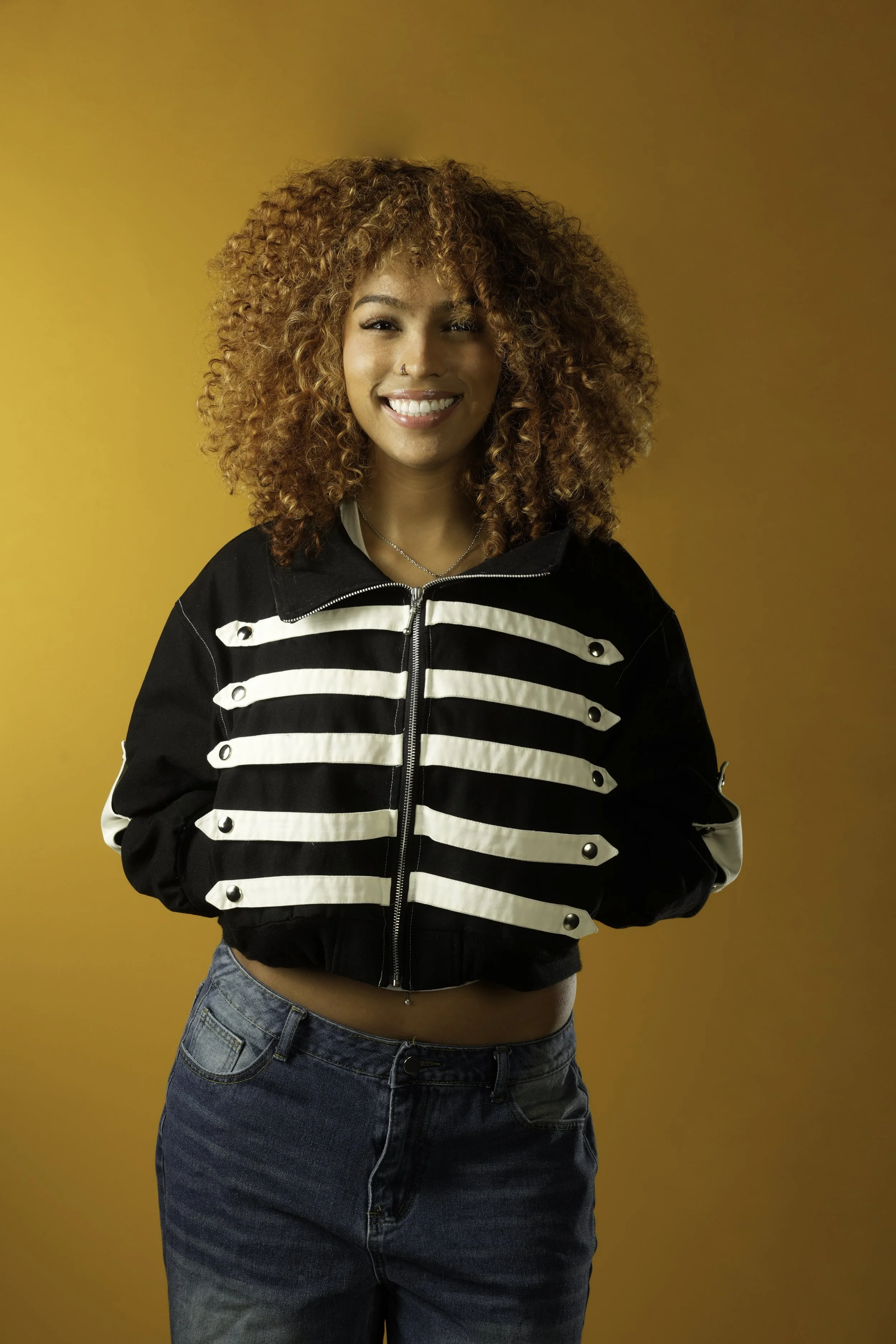 A young woman with curly red hair smiling, wearing a black and white striped jacket, a nose ring, and jeans, standing against a plain yellow background.