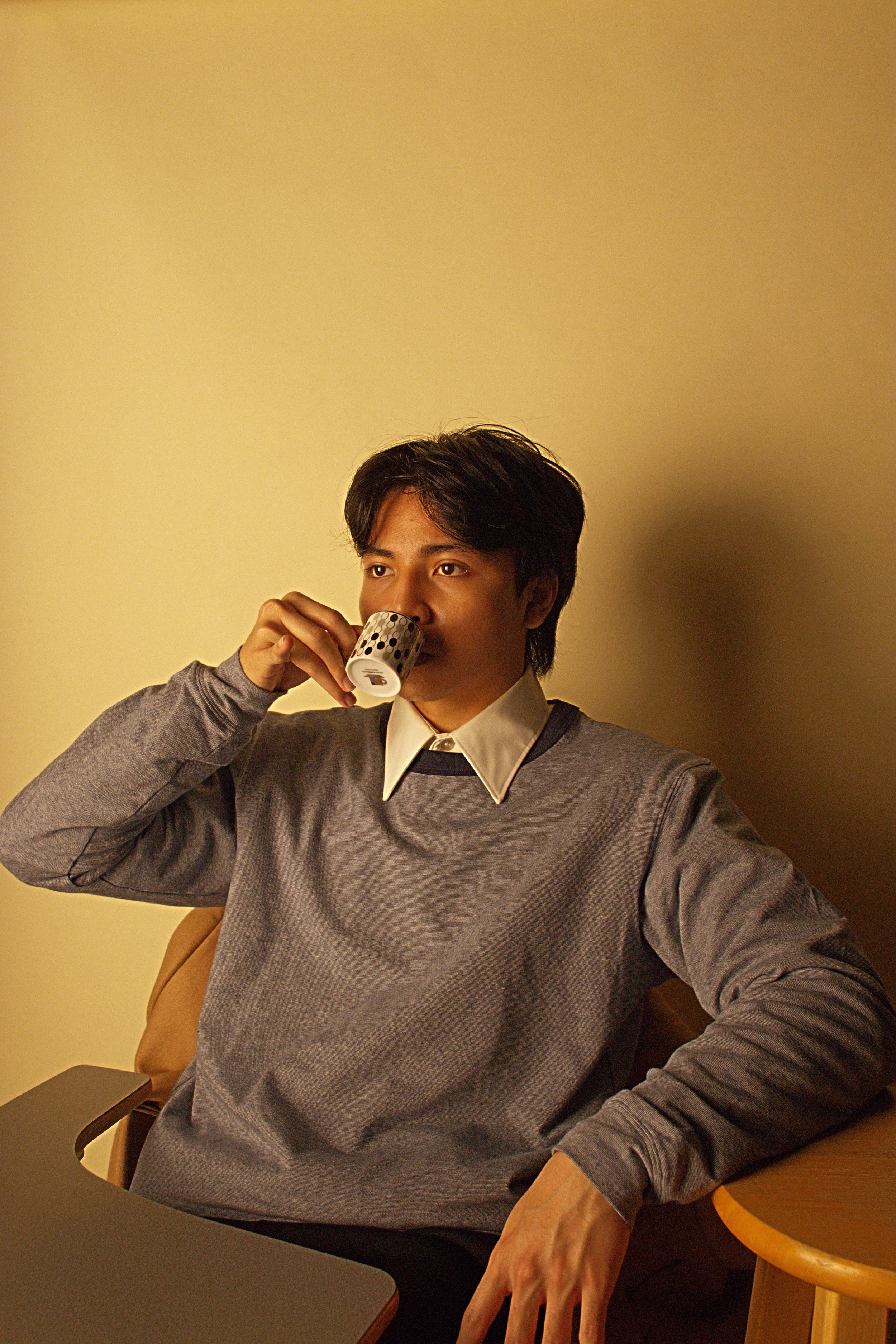 A young man with dark hair is sitting at a table, wearing a gray sweater over a white shirt and a black tie, drinking from a white mug with black dots. The background is a plain yellow wall.