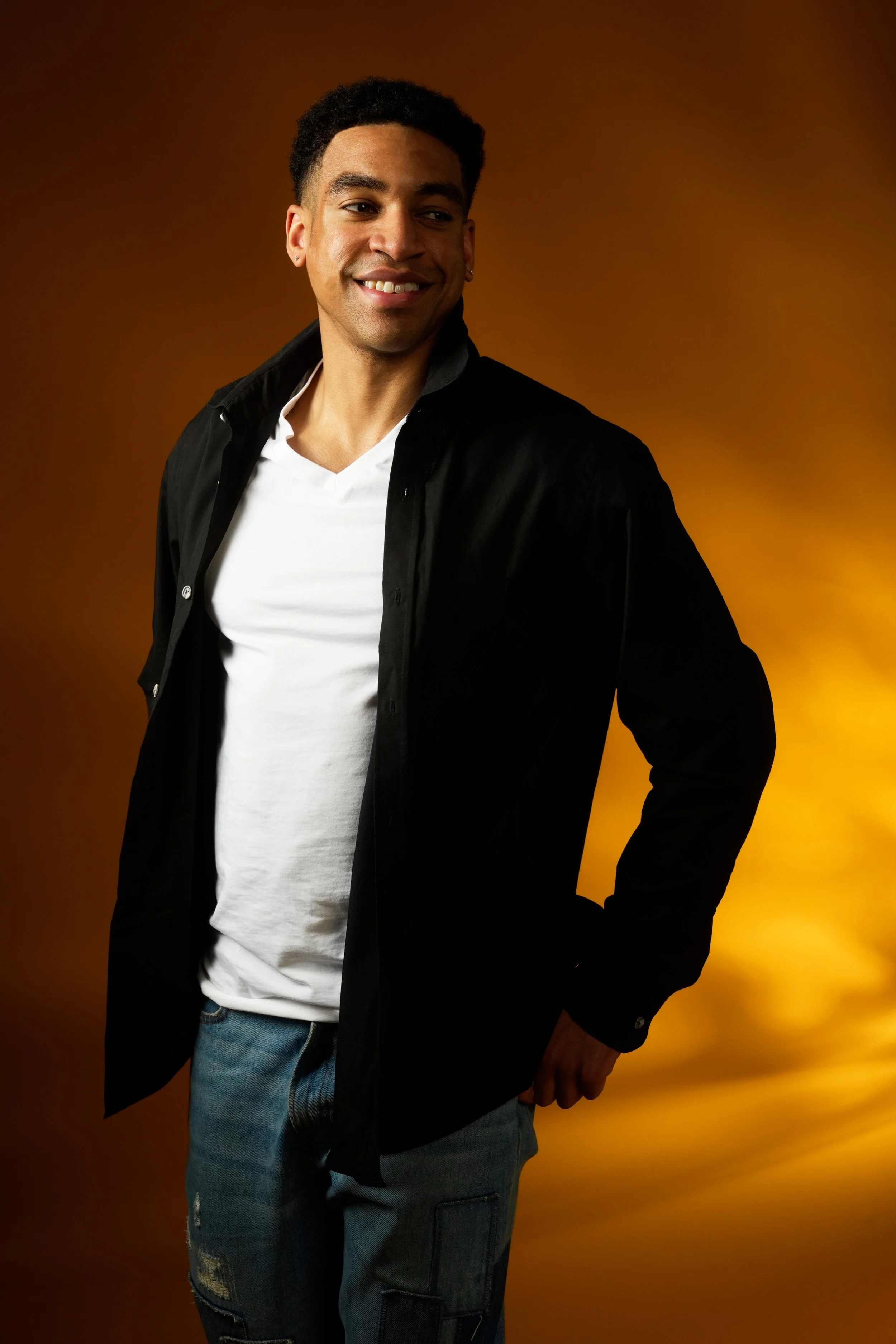 A young man with short curly hair, earrings, wearing a black jacket over a white t-shirt and jeans, smiling and standing against a warm gradient background.