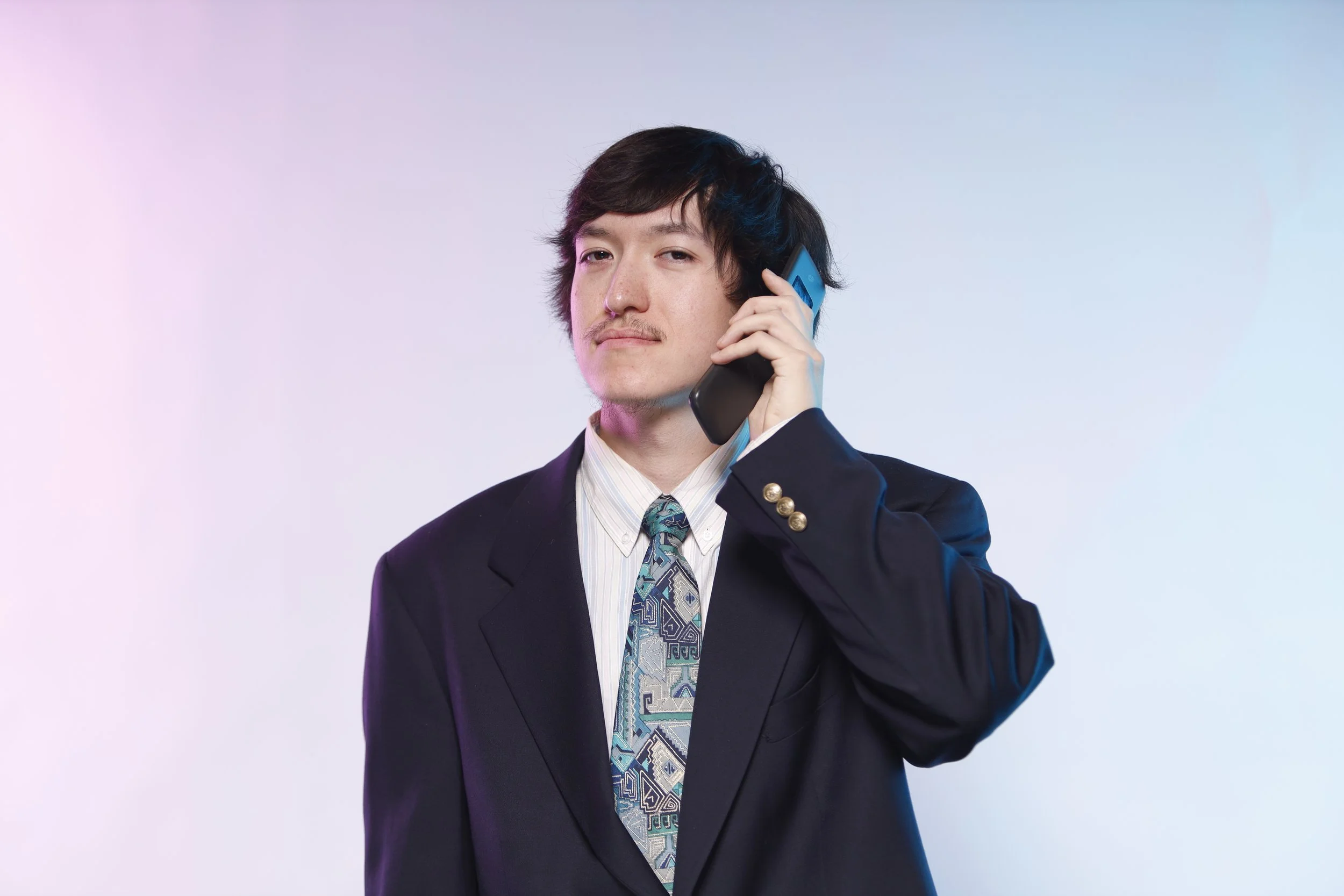 A man wearing a dark suit, white shirt, and patterned tie talking on a cell phone against a pastel-colored gradient background.